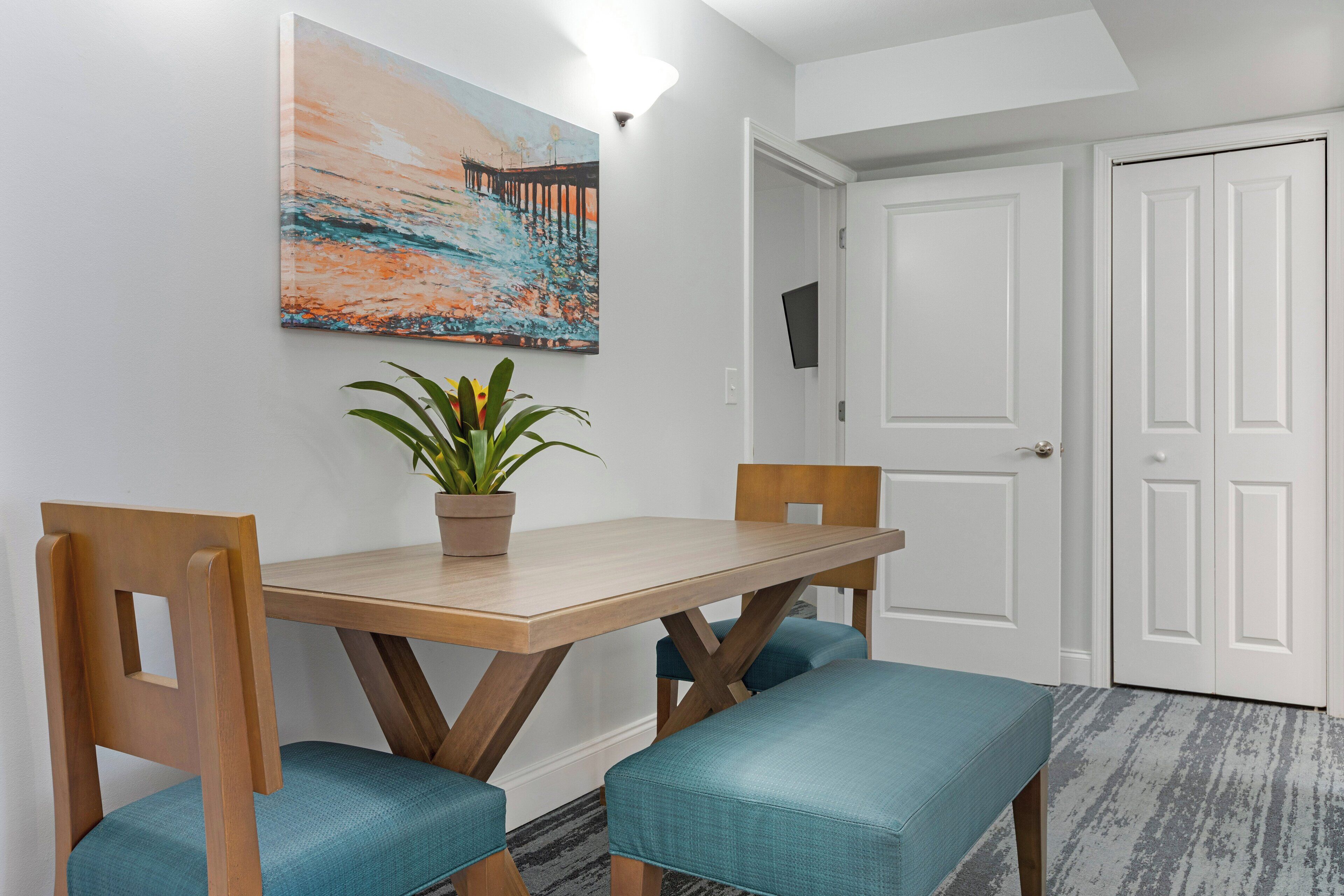A dining area with a wooden table and chairs, a potted plant, and a painting on the wall.