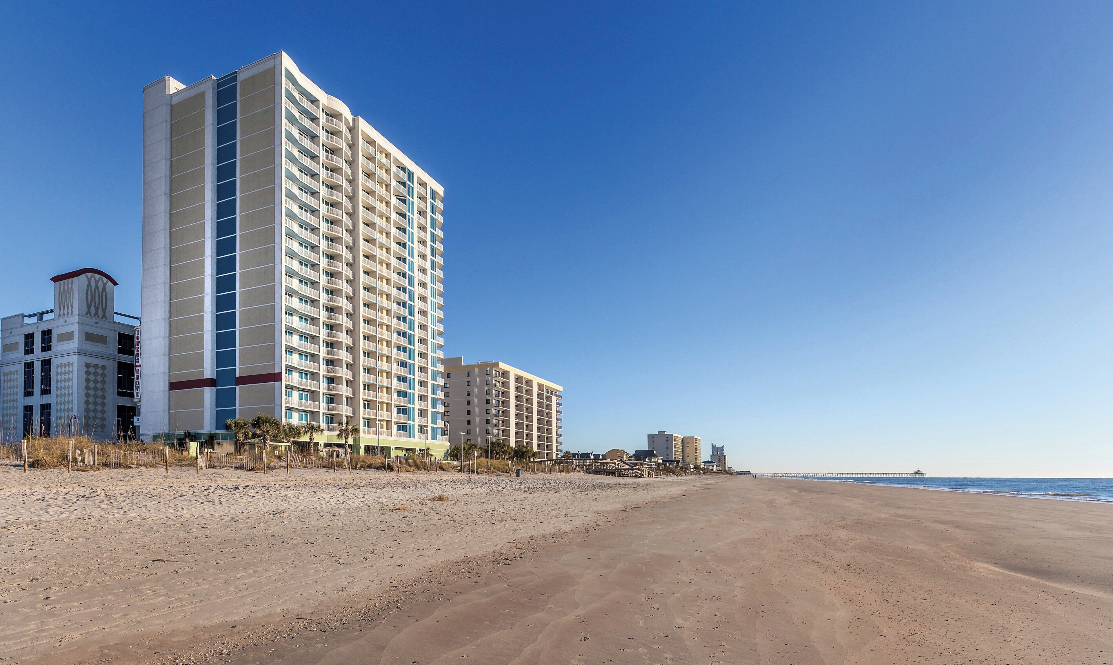Ein Hotel direkt am Strand mit mehreren Etagen, ein Sandstrand im Vordergrund und ein klarer, blauer Himmel.