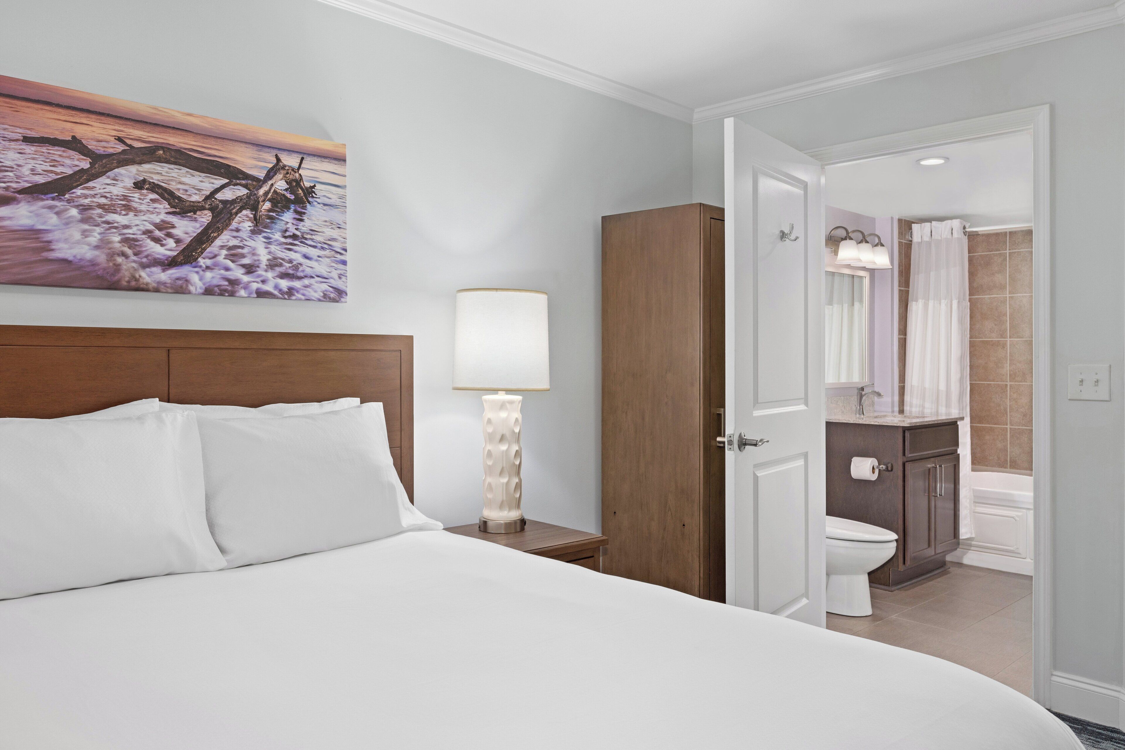 A hotel room with a bed, a nightstand with a lamp, a wooden wardrobe, and a bathroom visible through an open door.
