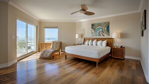 A bedroom with a large bed, a ceiling fan, a chair, a nightstand, and a view of the ocean.