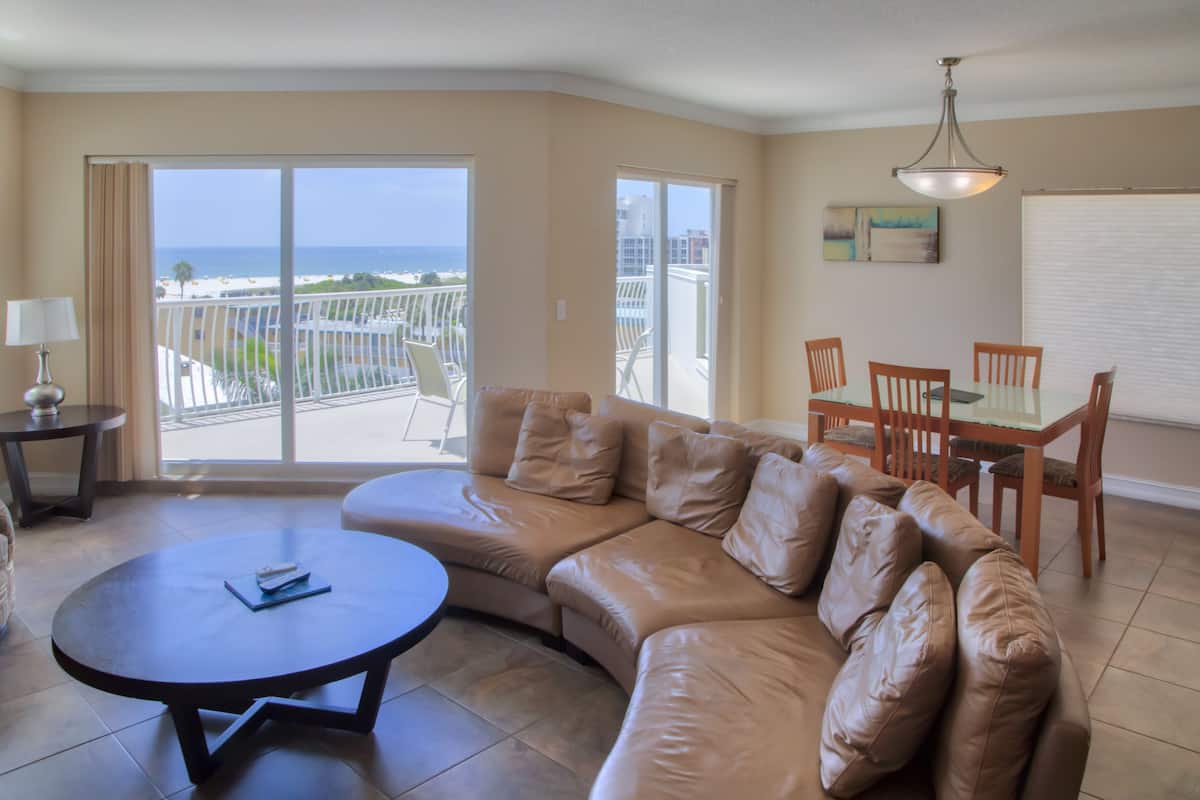 Een woonkamer met een beige leren bank, een ronde salontafel en een eettafel met stoelen. Er is een balkon met uitzicht op het strand en palmbomen.