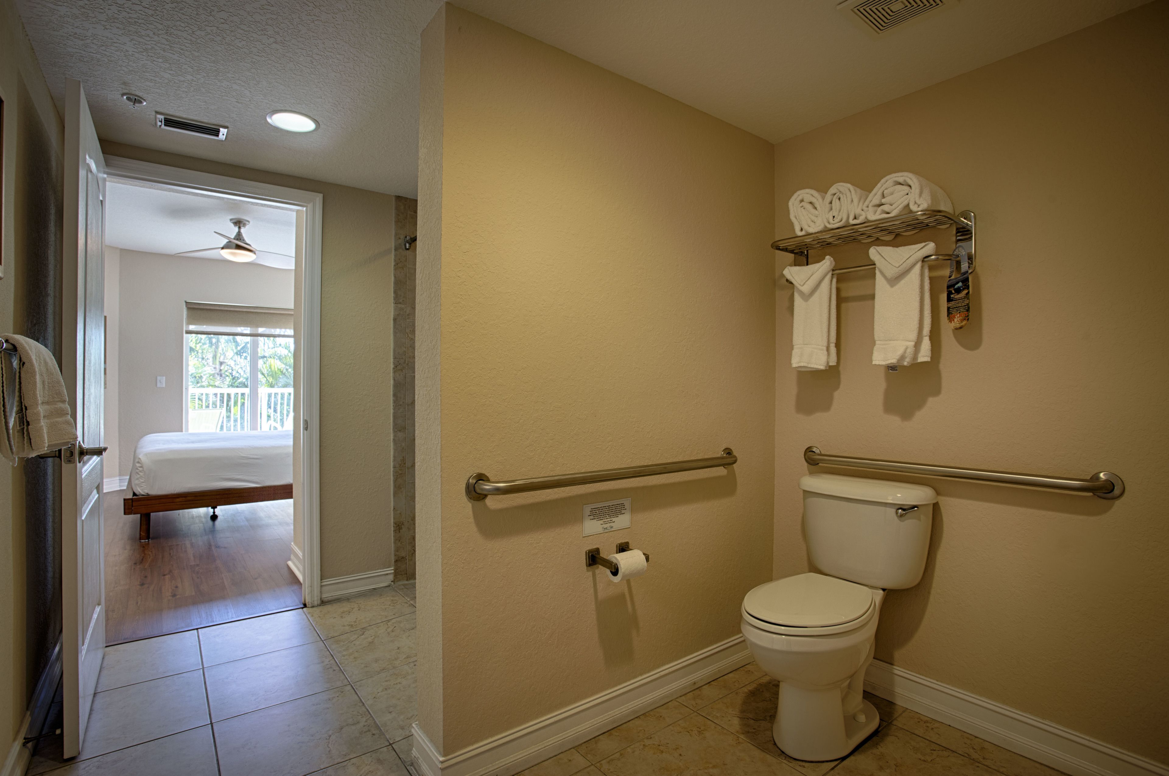 Une salle de bain équipée de toilettes, de barres d’appui, d’un porte-serviettes et d’une porte visible donnant sur une chambre.
