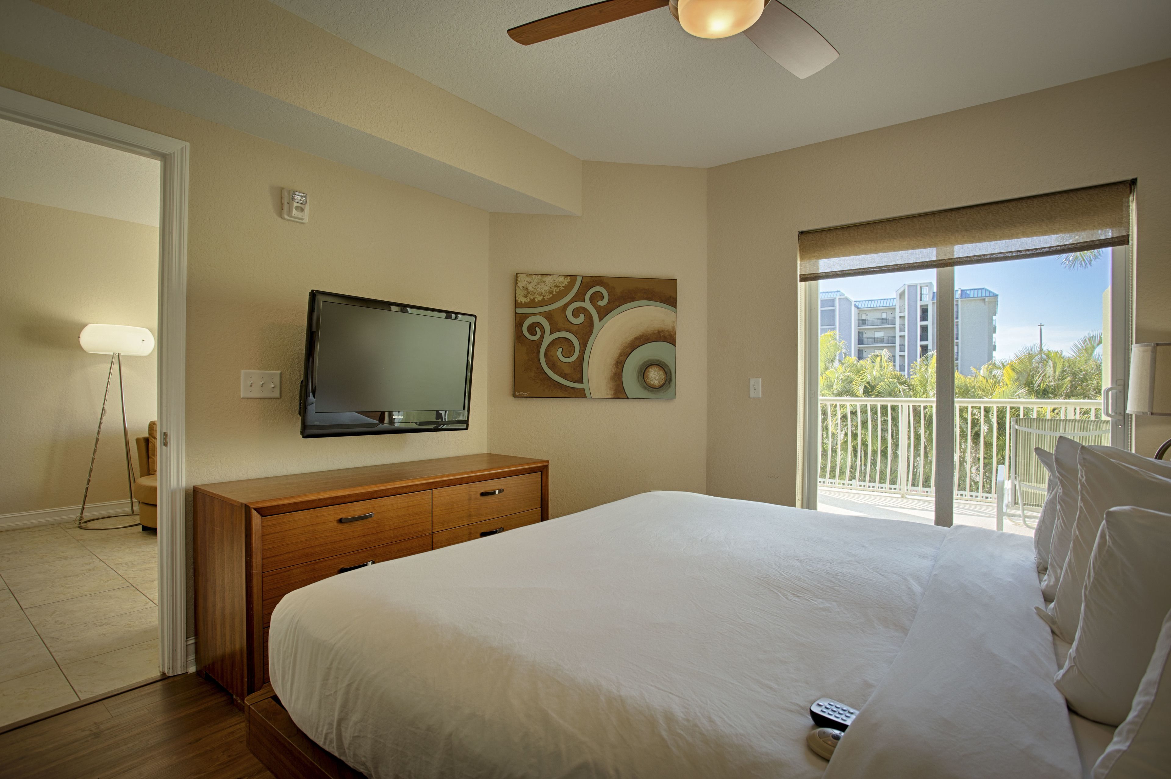 A hotel room with a large bed, a television mounted on the wall, a painting, and a balcony with a view of buildings.