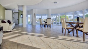 A spacious living area with a modern white sofa, a dining table with chairs, and large windows offering a view of the outdoors.