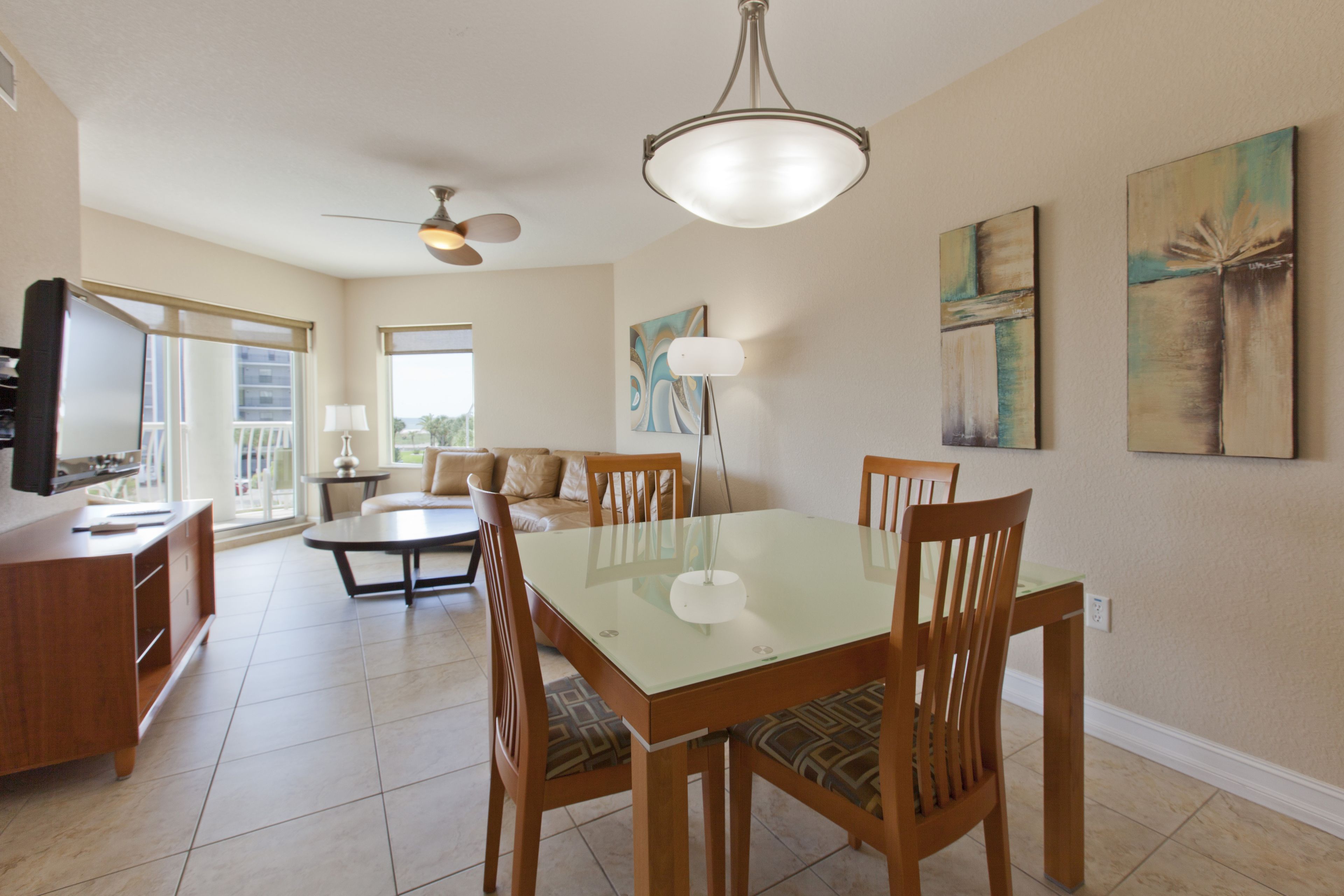 A spacious dining area with a glass-top table surrounded by wooden chairs, a ceiling fan, and artwork on the walls.