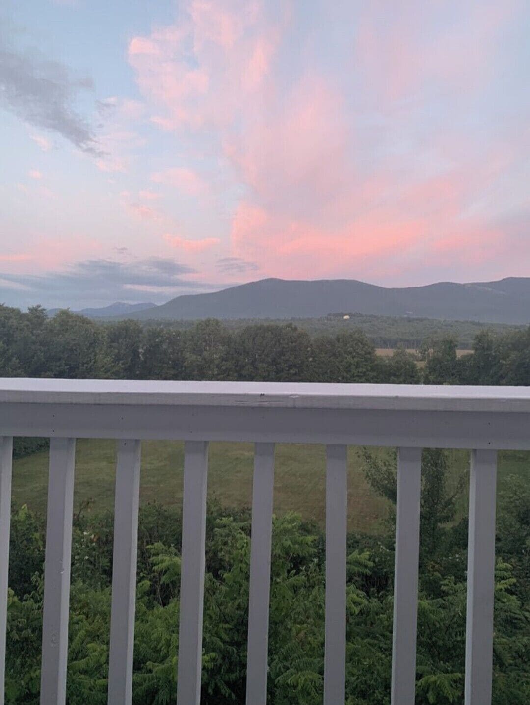 Un balcon doté d’une balustrade blanche, d’où l’on peut admirer un paysage composé d’arbres et de montagnes lointaines, sous un ciel rose et bleu.