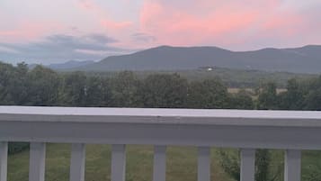 Ein Balkon mit weißer Brüstung, der auf eine Landschaft mit Bäumen und fernen Bergen unter einem rosa-blauen Himmel blickt.