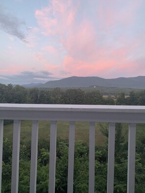 A balcony with a white railing overlooking a landscape with trees and distant mountains under a pink and blue sky. - North Conway Mountain Inn (North Conway)