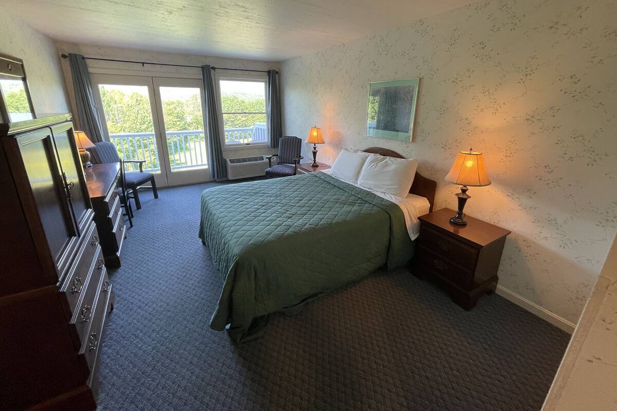 A hotel room with a bed, two bedside lamps, a chair, a desk, and a view of the outdoors through a sliding door.