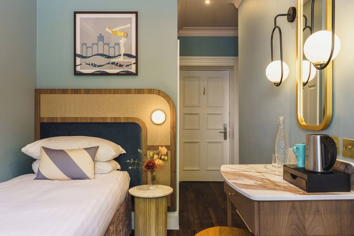Quarto de hotel com cama, mesa de cabeceira e banheiro com espelho, pia e iluminação.