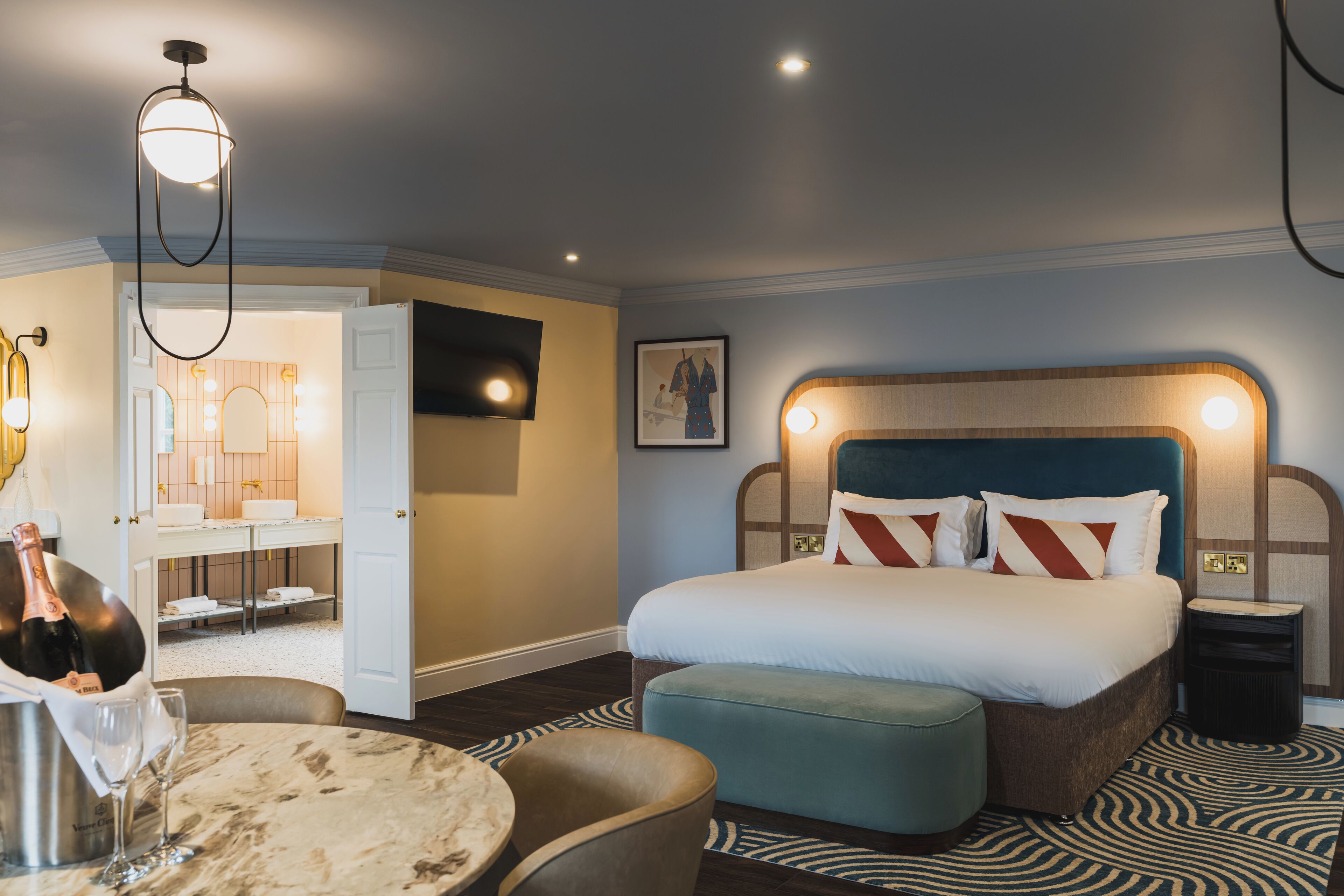 A hotel room with a large bed, a sitting area with a table and chairs, a minibar, and a bathroom visible through an open door.