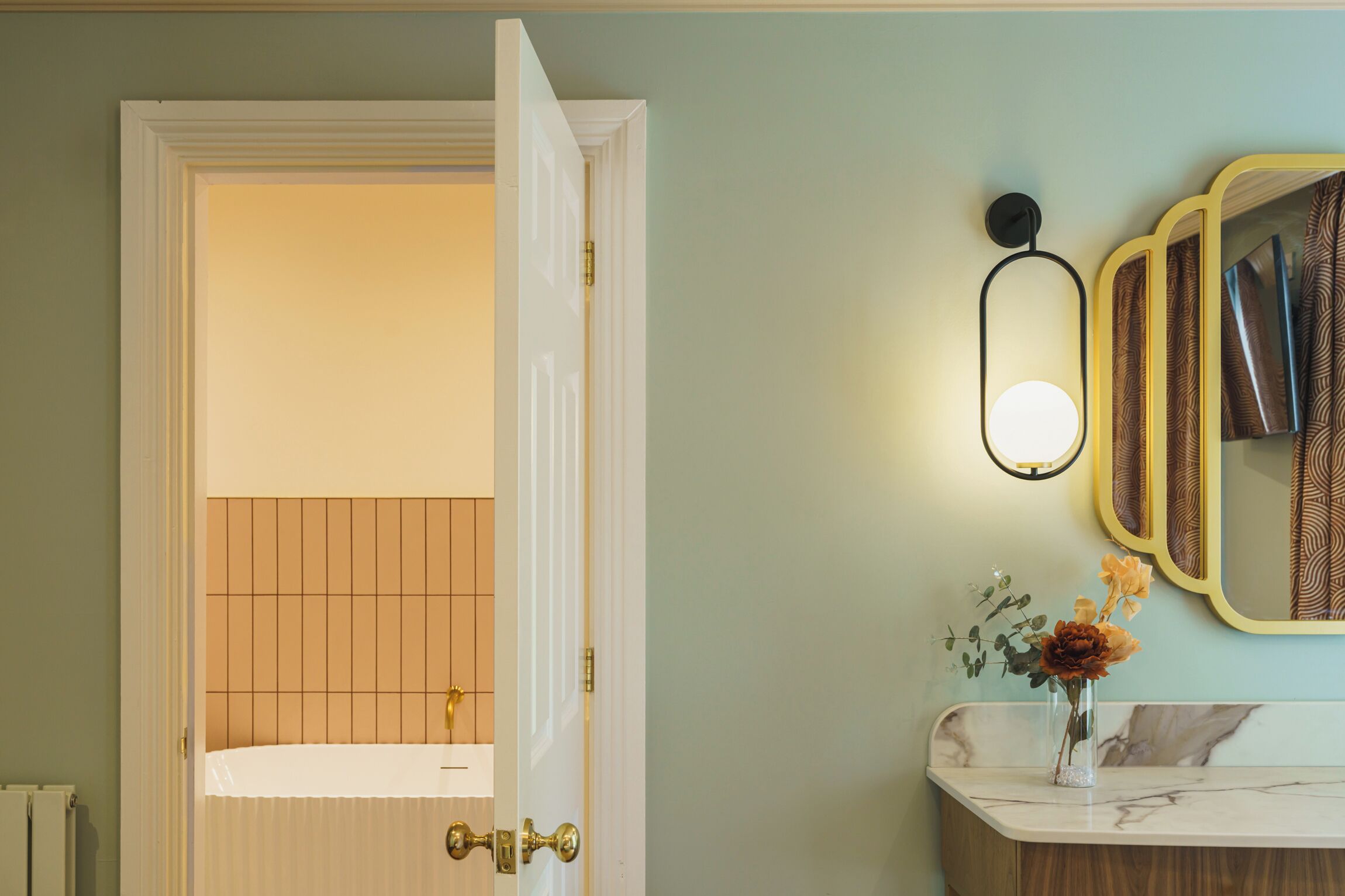 A bathroom with a white bathtub, a door leading to a room, a wall-mounted mirror with a patterned frame, and a vase with flowers on the counter.