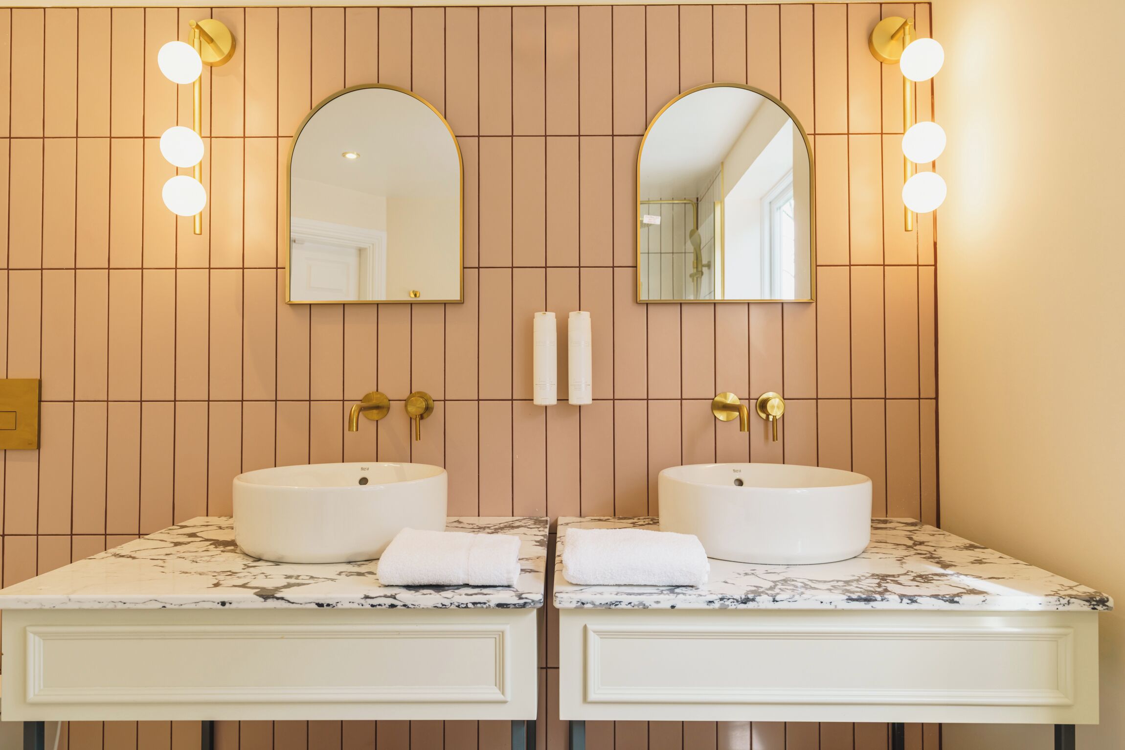 A bathroom with two sinks, each with a white marble countertop and a gold faucet. There are two wall-mounted mirrors with arched tops and a light fixture above each mirror.
