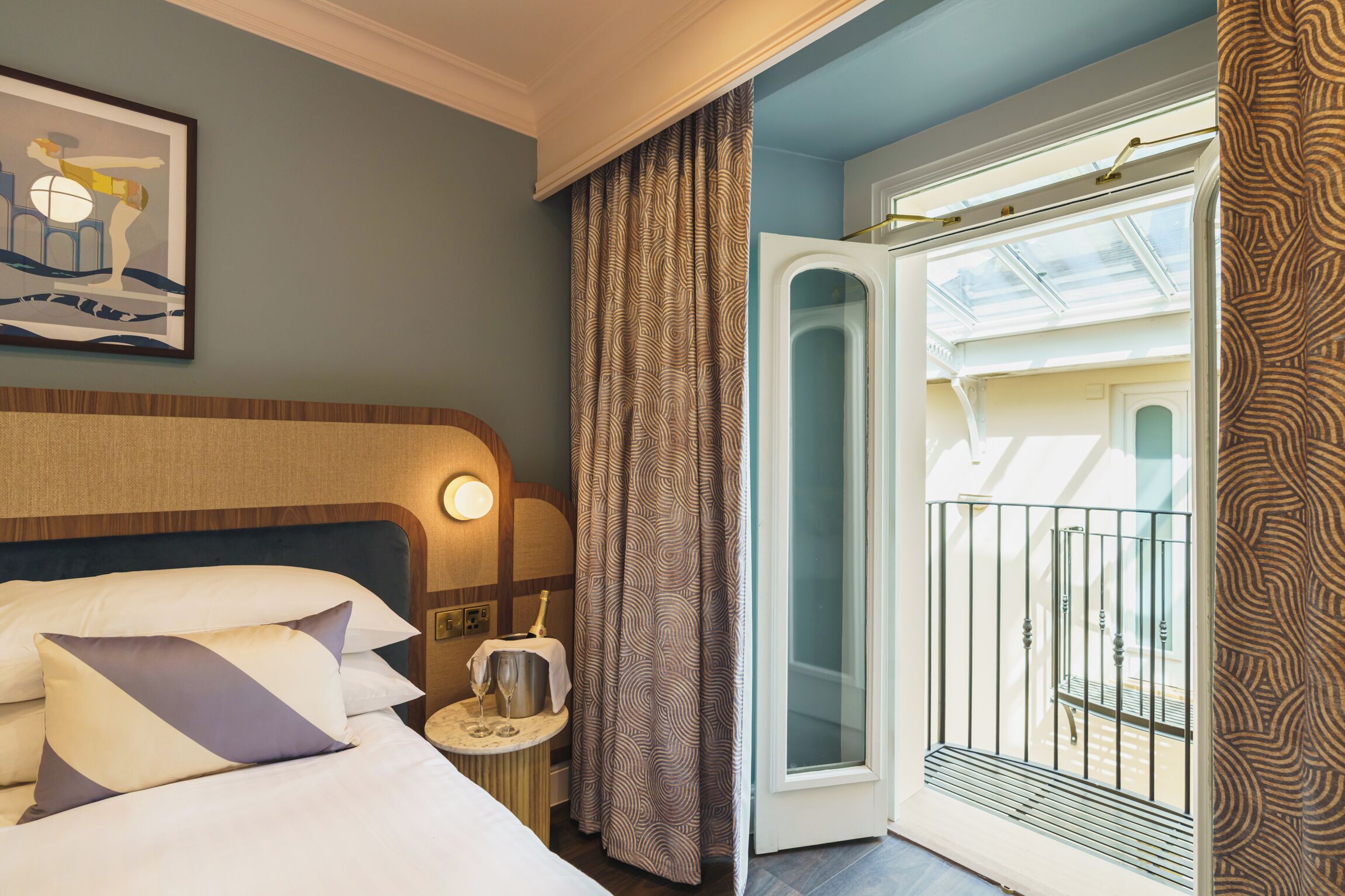 A hotel room with a bed, a nightstand, a glass-enclosed balcony, and a patterned curtain.