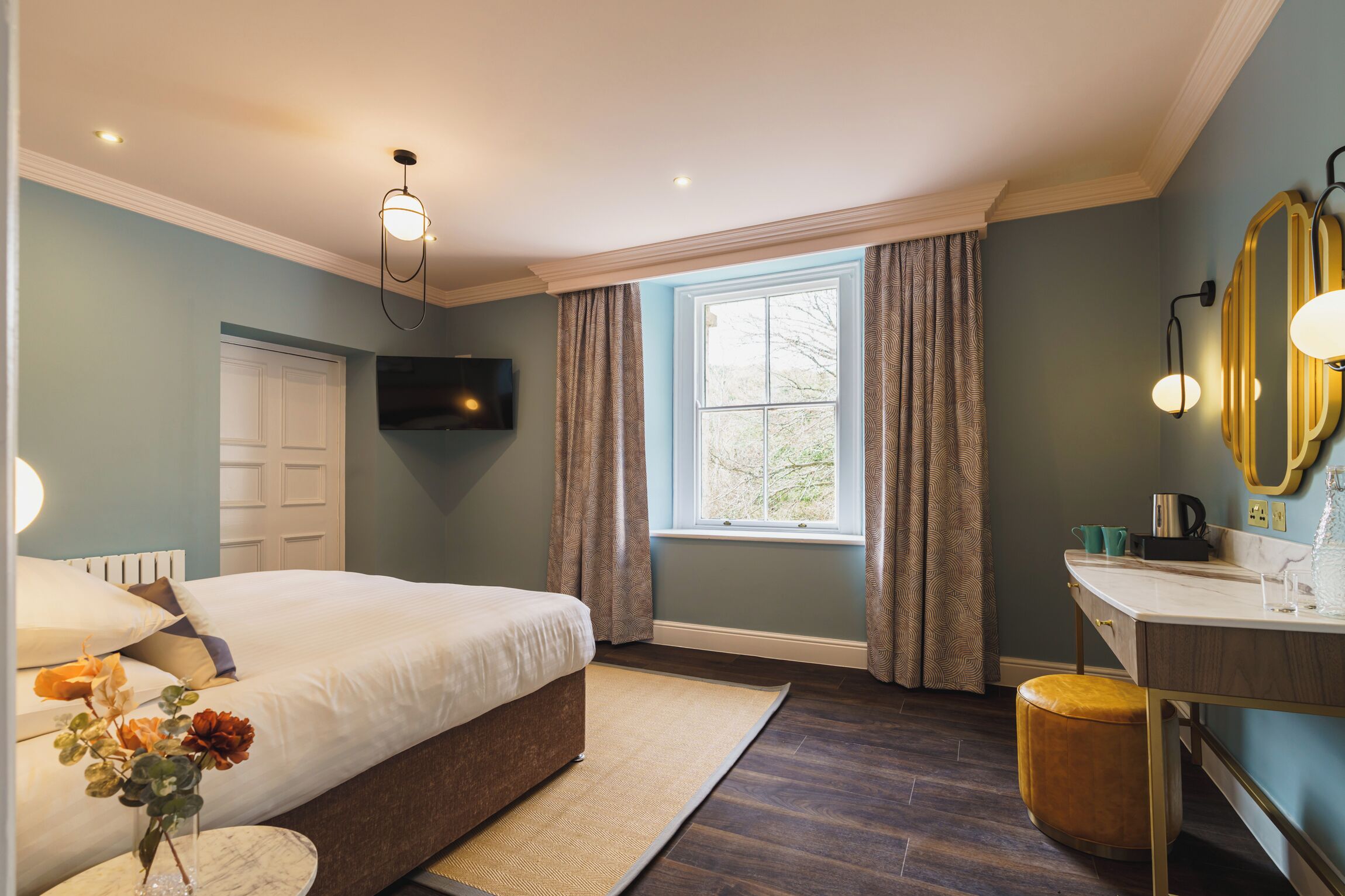 A bedroom with a bed, a bedside table, a window with curtains, a flat-screen TV, and a bathroom with a sink and mirror.