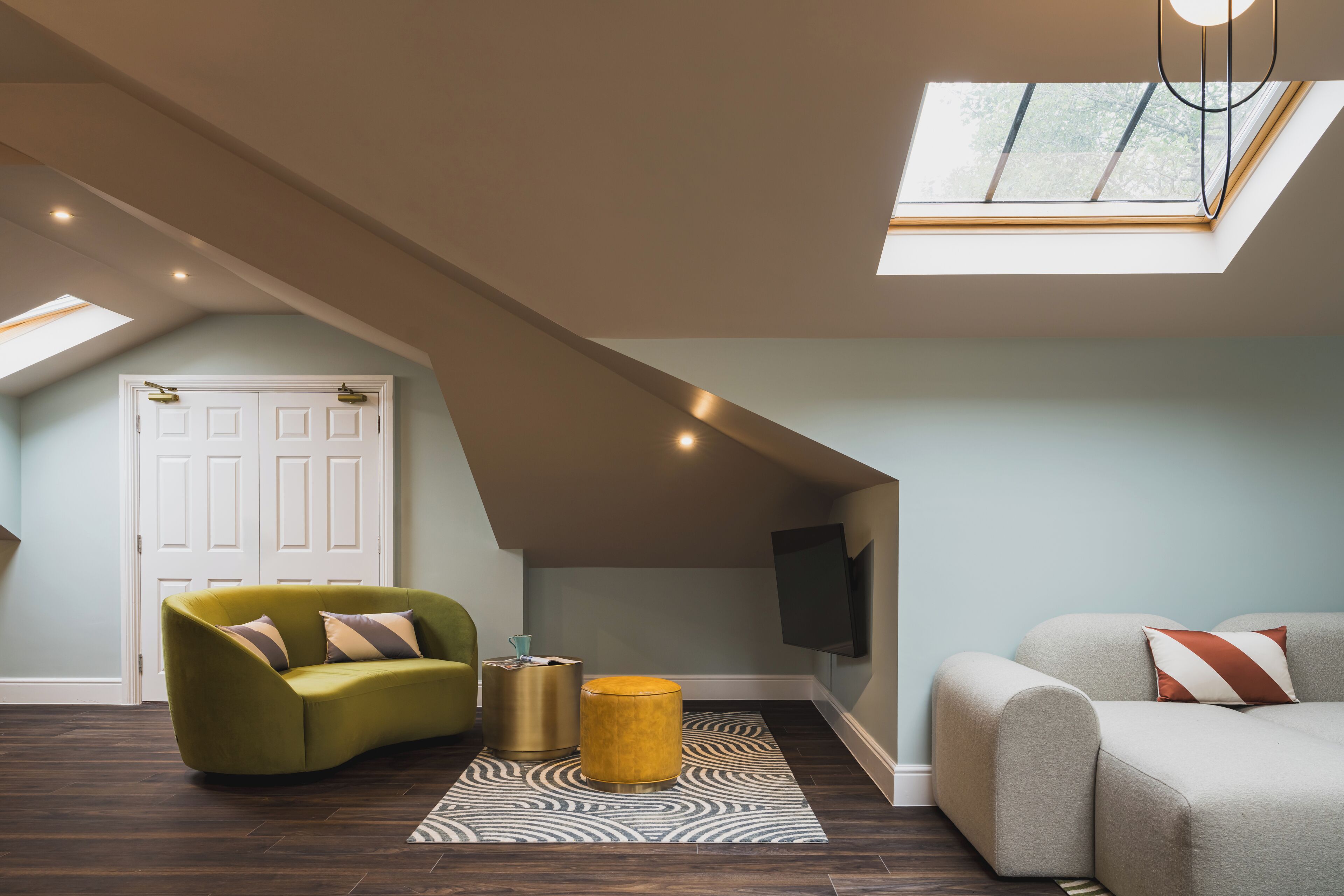 Een loft-achtige kamer met een schuine plafond, groot raam, flatscreen televisie aan de muur en twee banken.