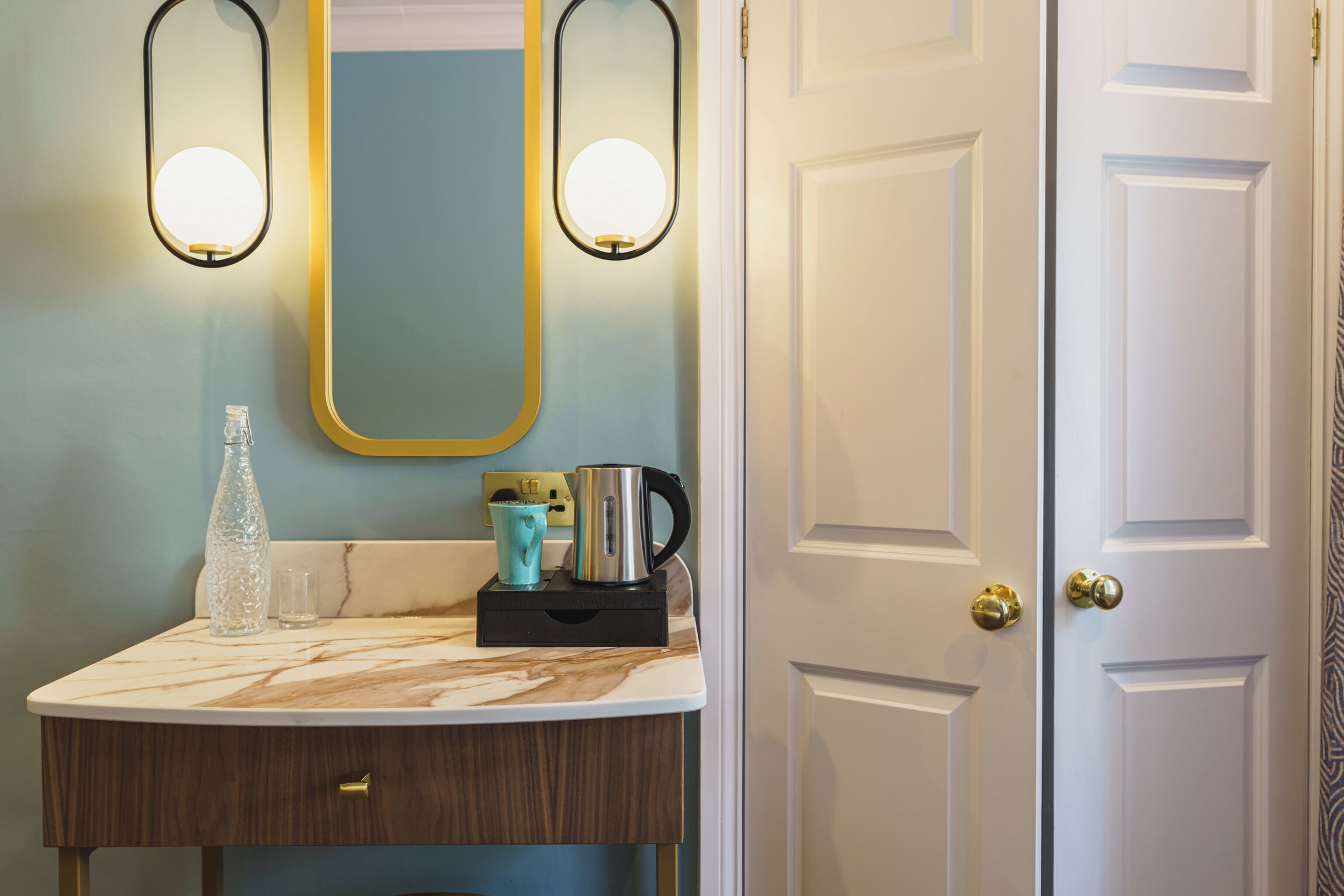 Baño con encimera de mármol, una botella de vidrio, taza azul y cafetera. Hay dos luces empotradas y una puerta con diseño decorativo.