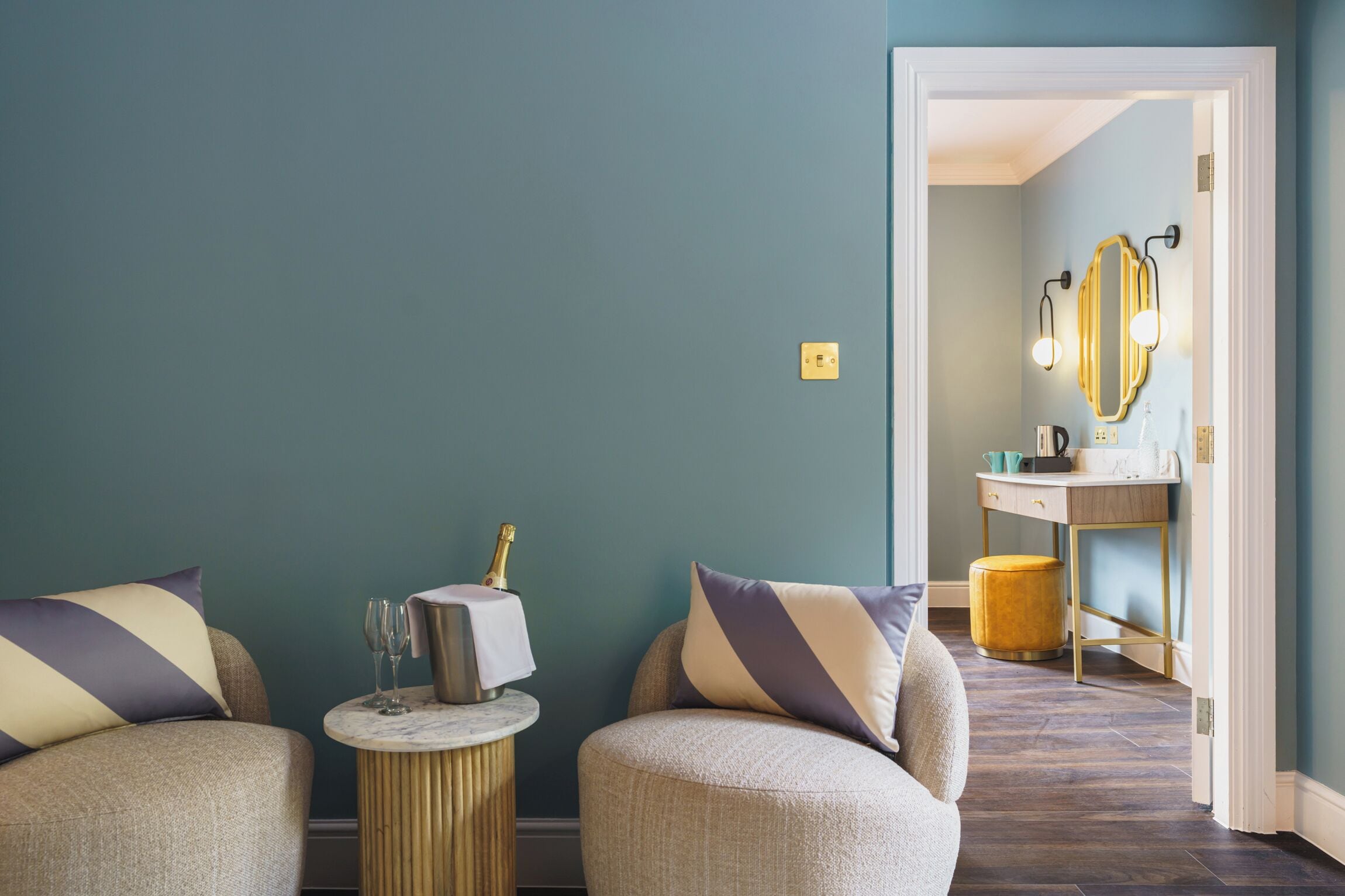 Une pièce aux murs de couleur turquoise, une table ronde avec une bouteille de champagne et des verres, deux fauteuils avec des coussins à motifs, et un miroir avec un cadre doré placé au-dessus d’une coiffeuse.
