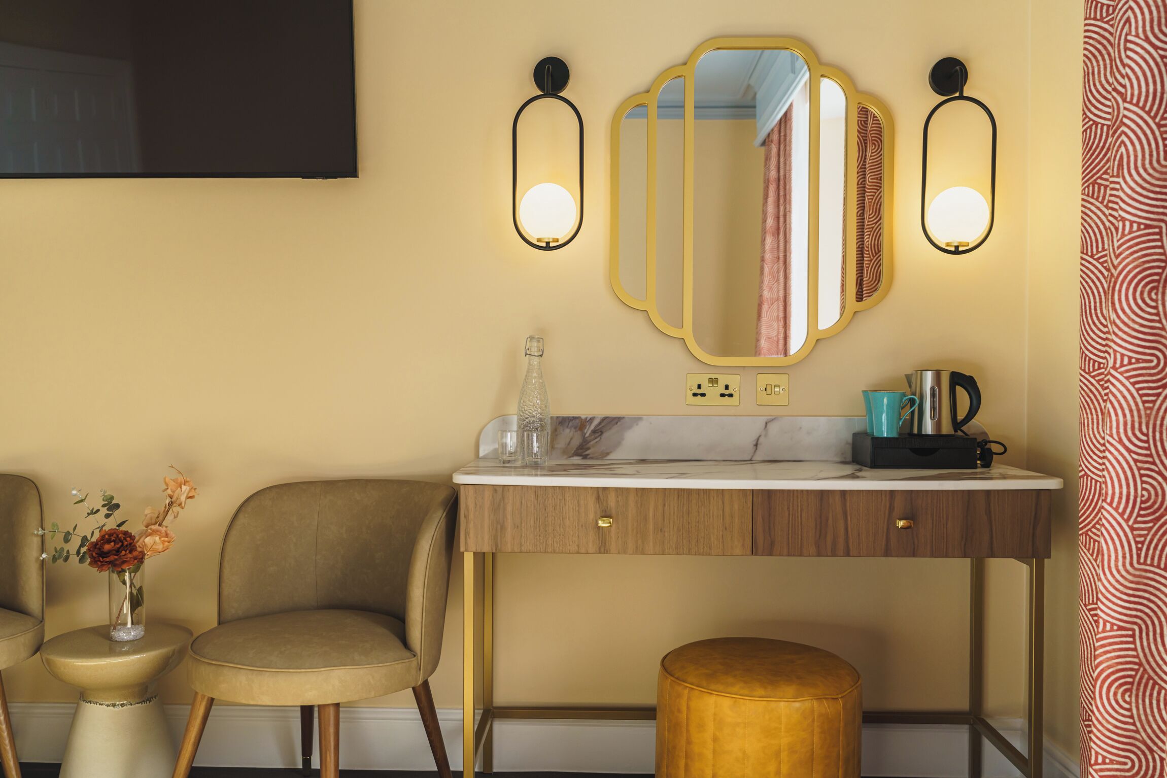 A hotel room with a vanity, two wall-mounted mirrors, a vase with flowers, a coffee maker, and a small round stool.