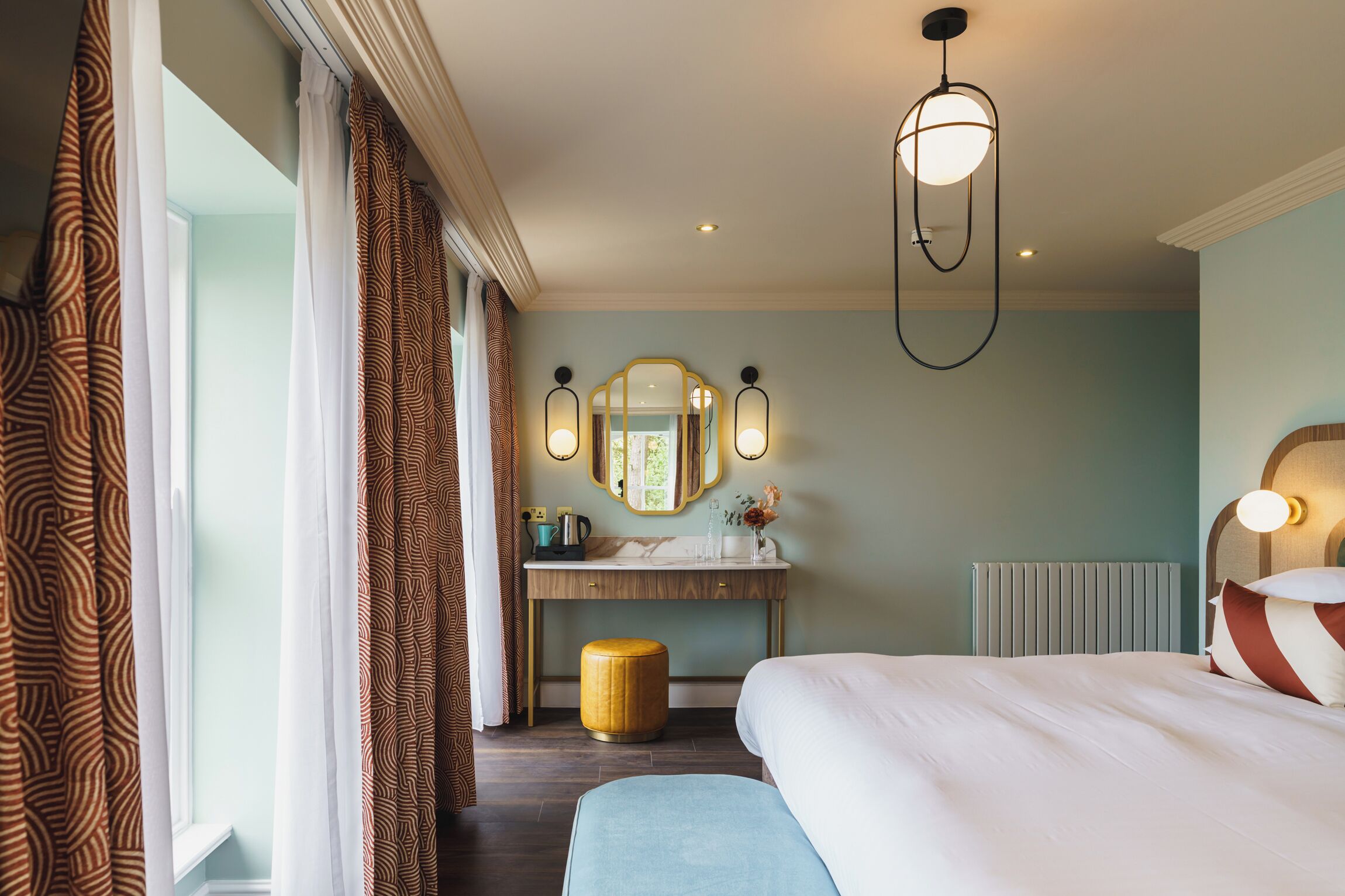 A hotel room with a bed, a vanity with a mirror, a stool, and a radiator.