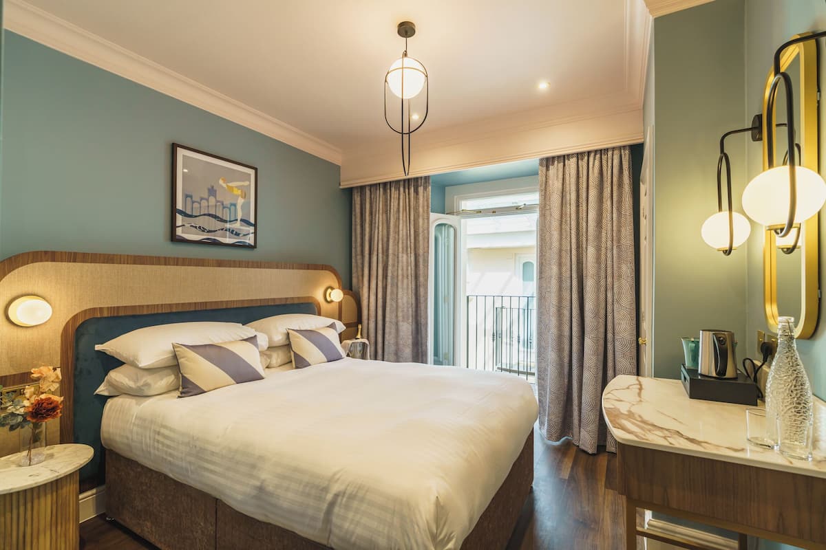 Quarto de hotel com uma cama grande, uma mesa de cabeceira com luminária, espelho e vista para a varanda.