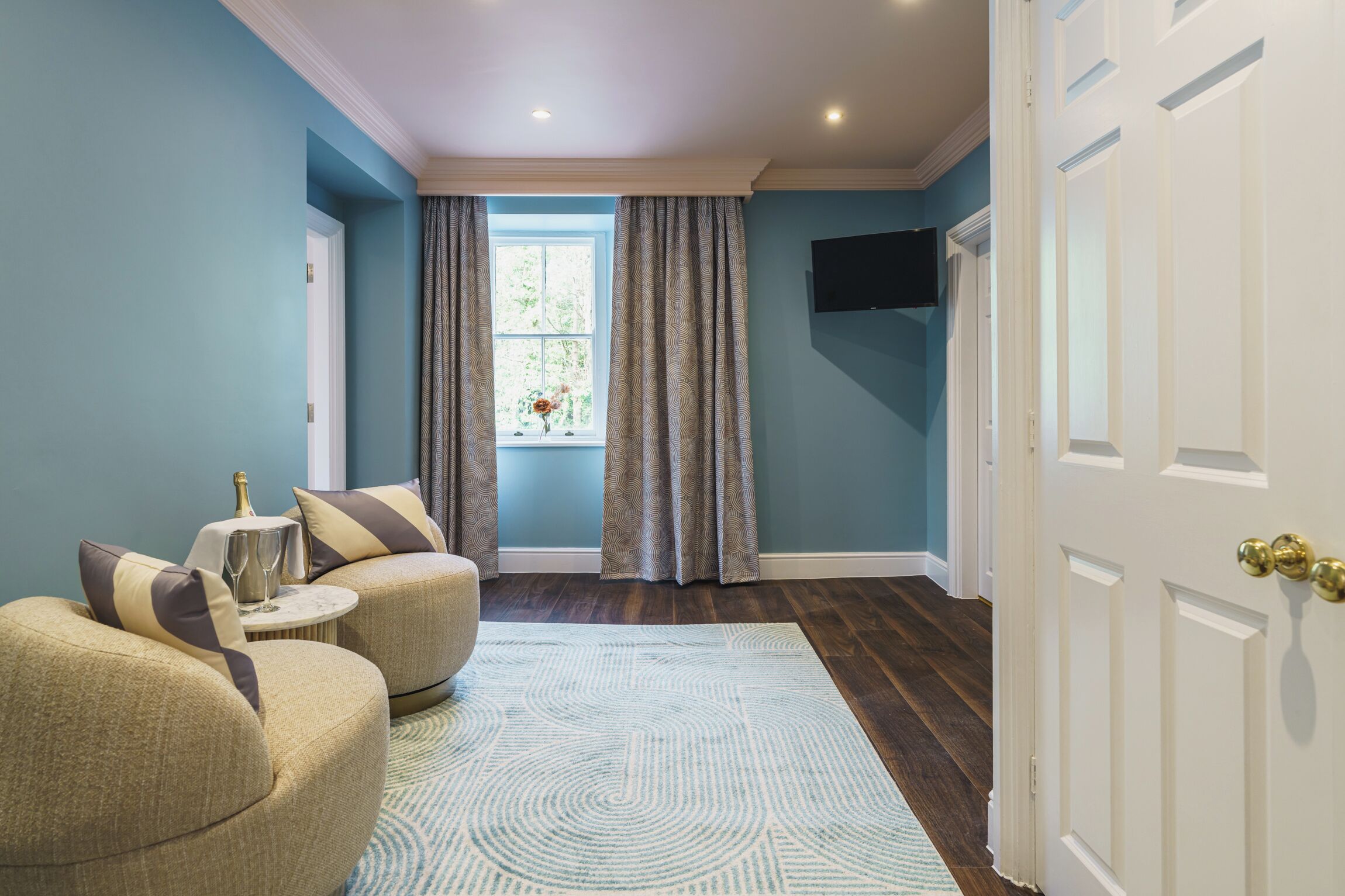 A room with a blue wall, a white door, and a window with curtains. There is a round table with a chair and a small rug.