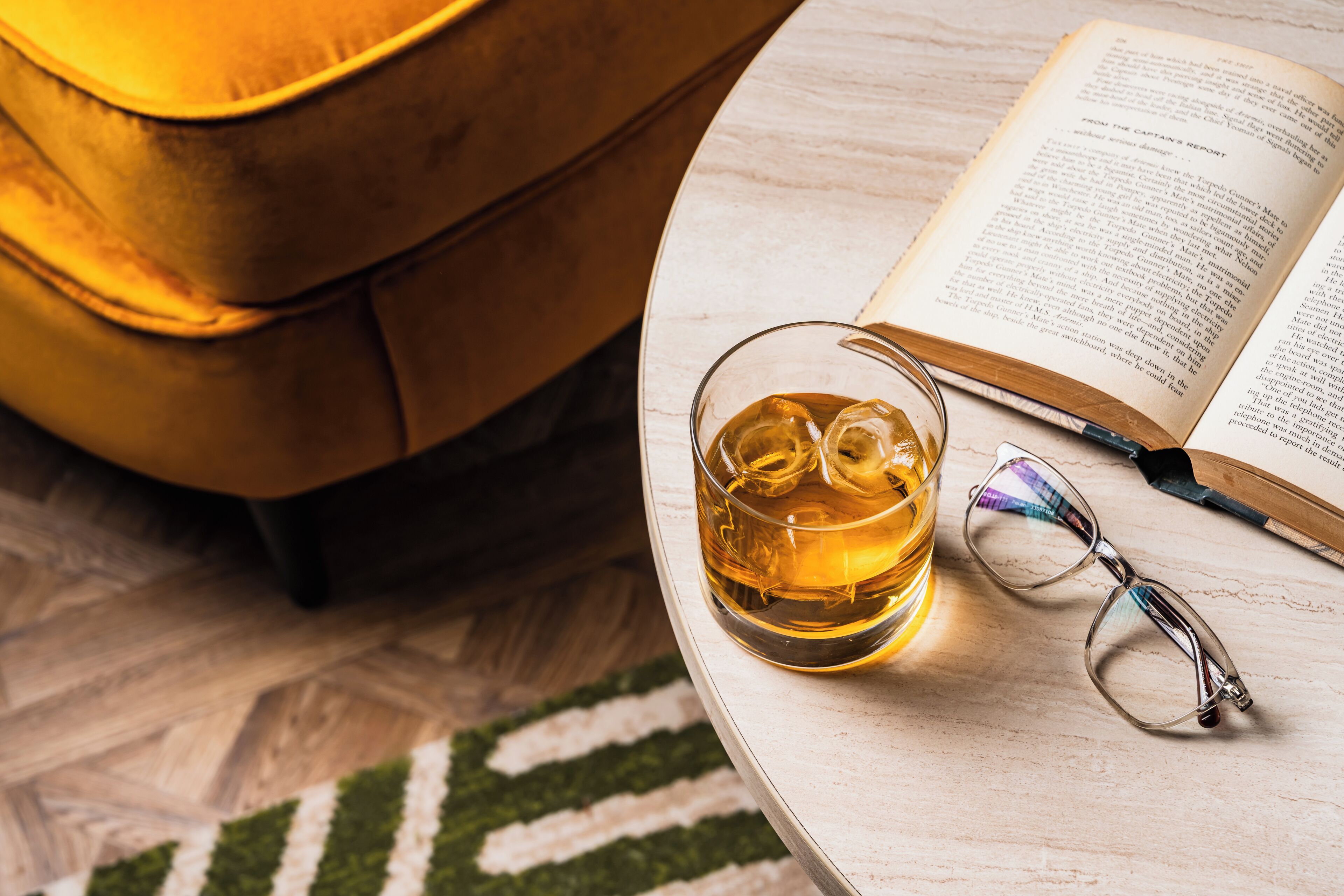 Une table ronde sur laquelle se trouvent un verre contenant un liquide ambré, une paire de lunettes et un livre ouvert.