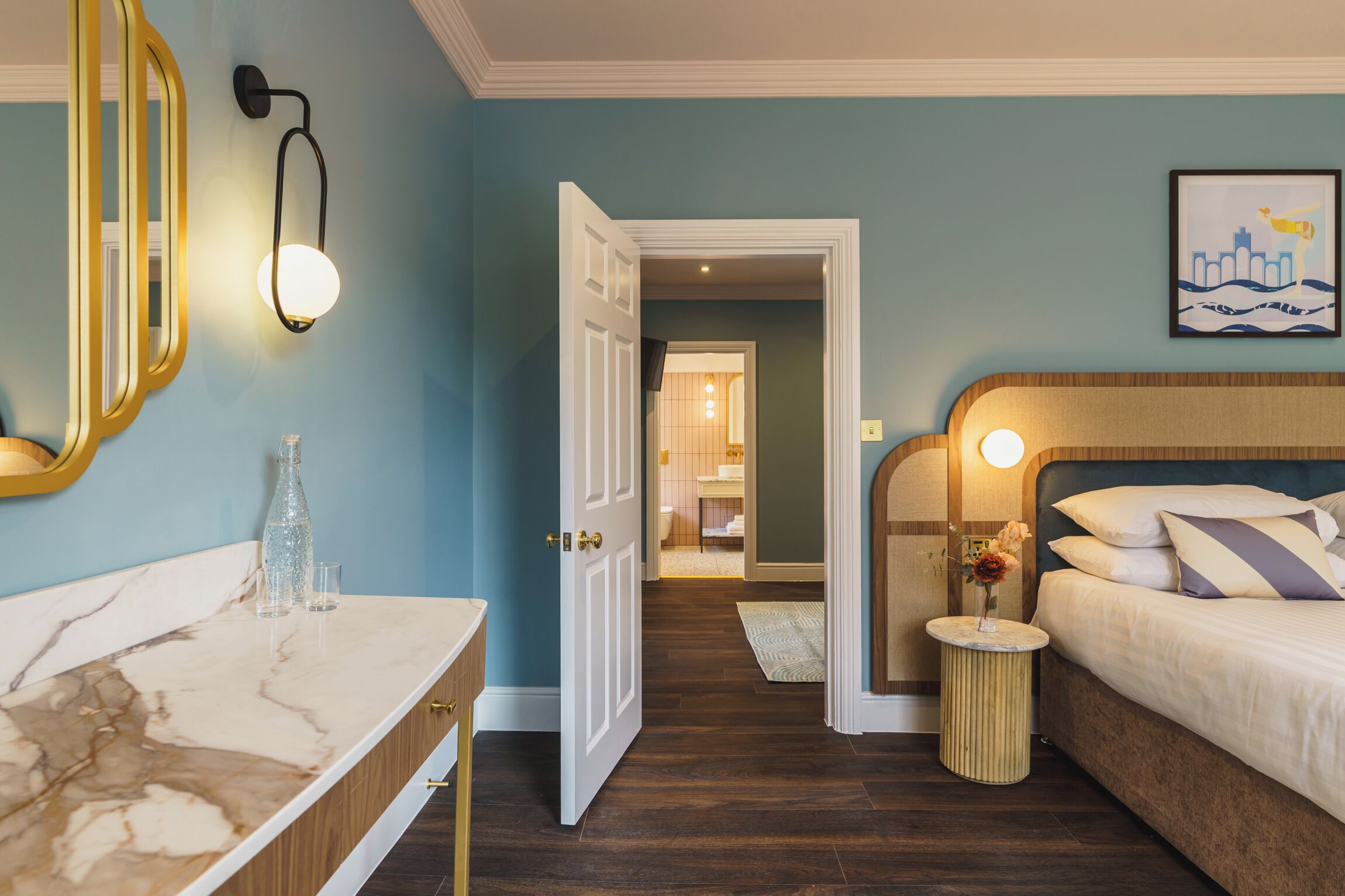 Une chambre d’hôtel avec un meuble-lavabo en marbre, un lit avec des oreillers à rayures, une table de chevet avec un vase et un tableau encadré au mur.