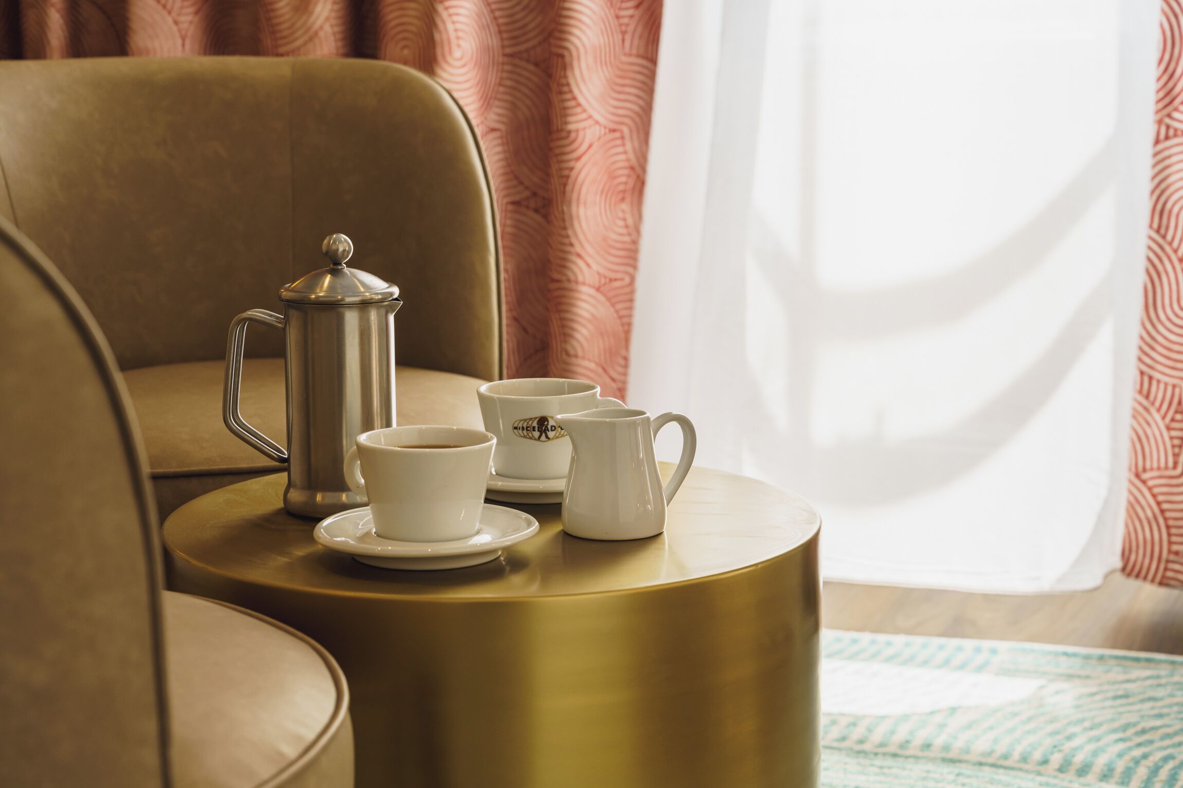 A round table with a coffee pot, two cups, and a jug, set on a gold-colored table in a room with a brown chair and patterned curtains.