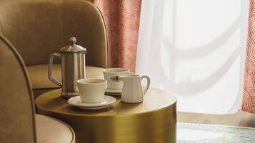 A round table with a coffee pot, two cups, and a jug, set on a gold-colored table in a room with a brown chair and patterned curtains.