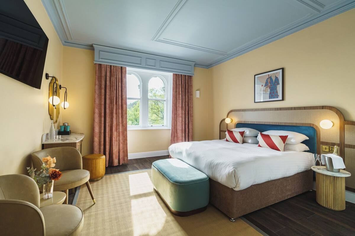 Quarto de hotel com uma cama grande, uma escrivaninha com abajur, uma cadeira, uma mesinha de centro com um vaso de flores e uma janela com cortinas.