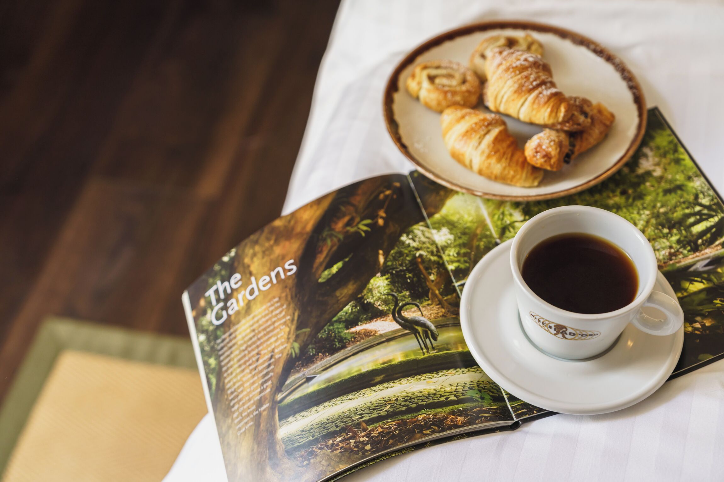 Una revista con una portada alusiva a la naturaleza, un plato con cruasanes y una taza de café sobre una mesa.