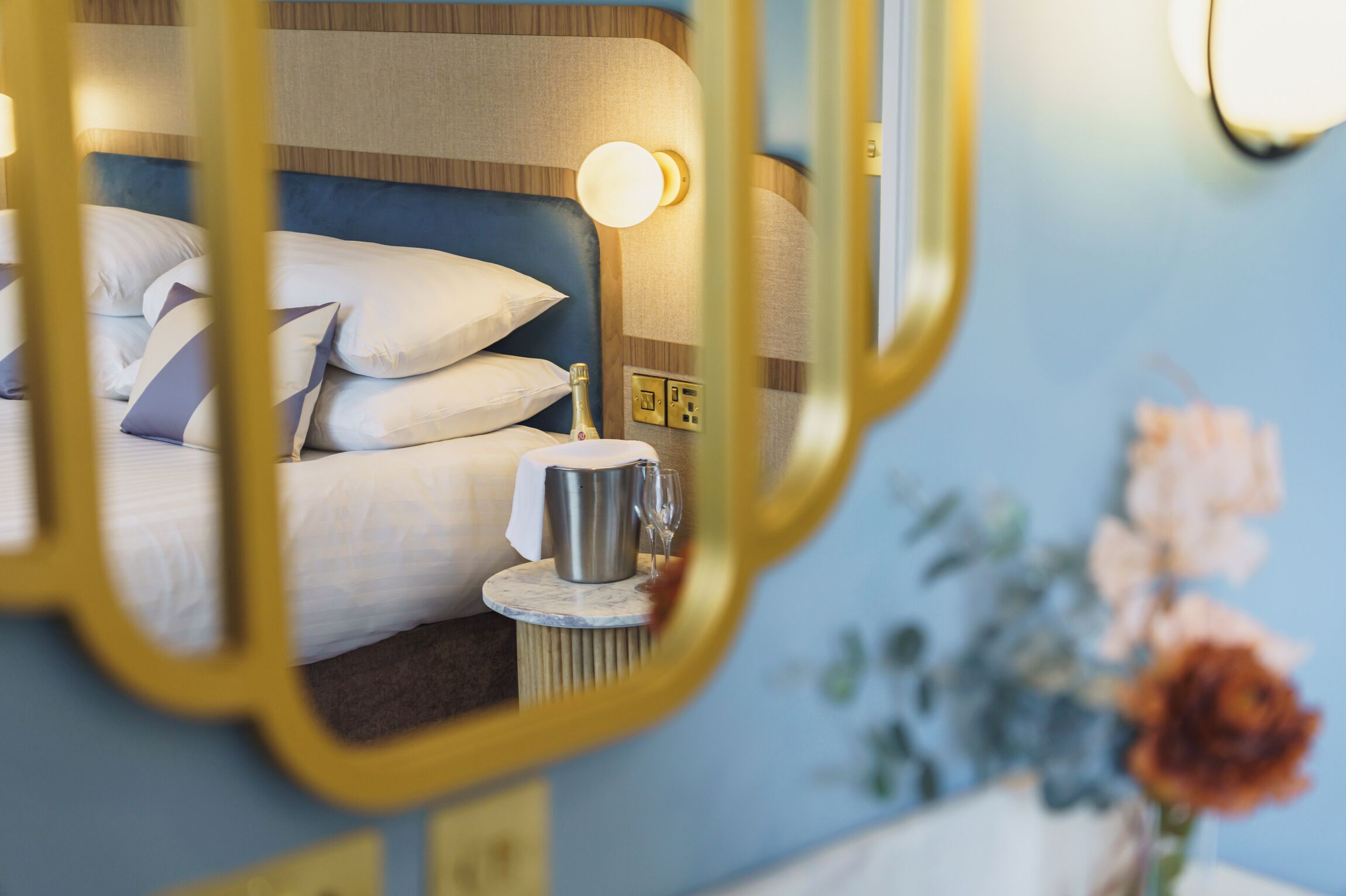 A hotel room with a bed, a bedside table with a bottle and glass, and a mirror with a gold frame.