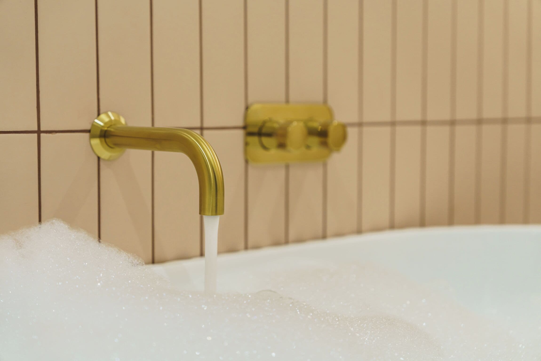Una bañera con agua corriente y burbujas, un grifo dorado y un panel de control amarillo en una pared azulejada.