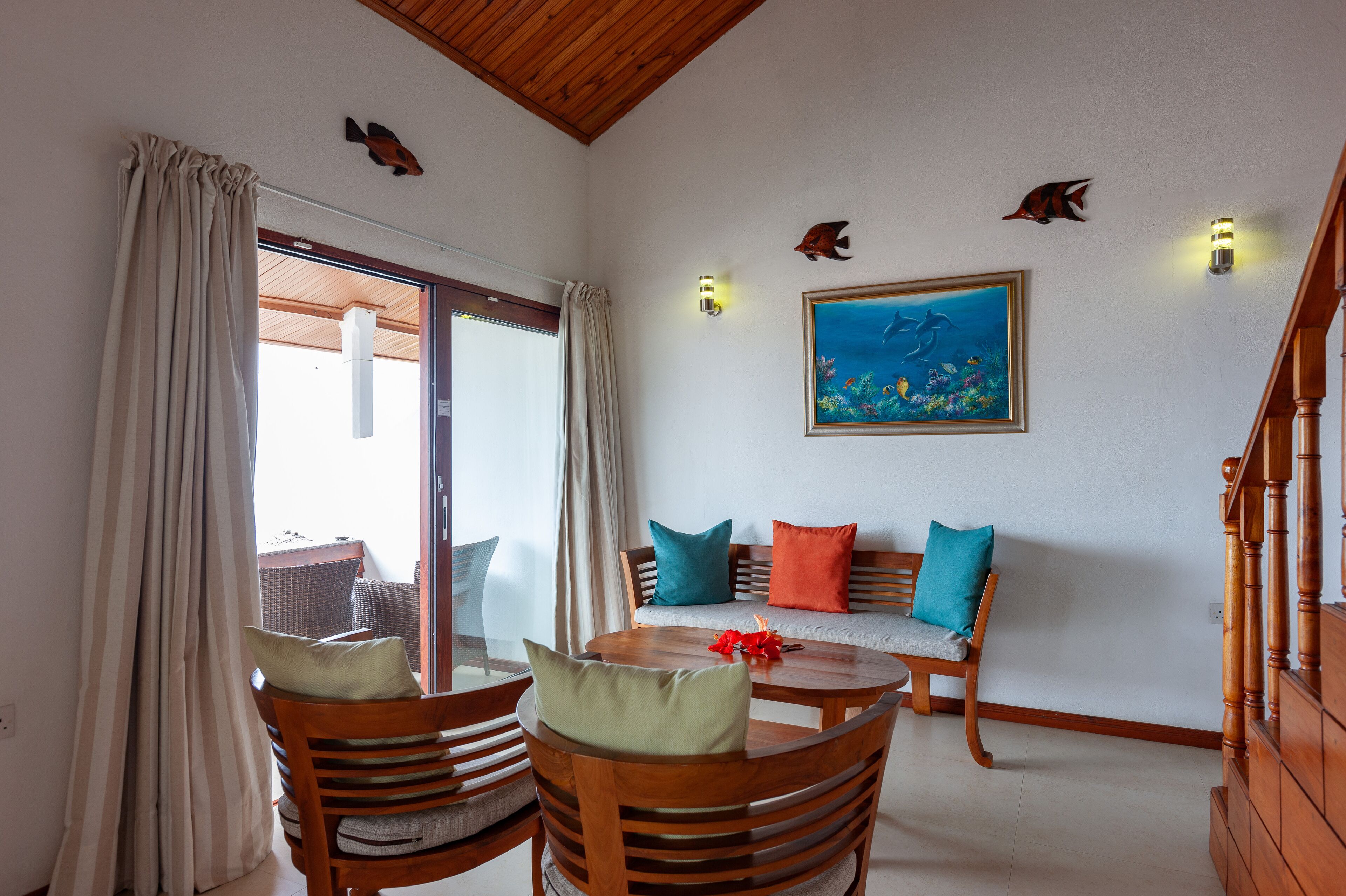 Una habitación con una mesa de madera, sillas, un sofá con cojines de colores, un cuadro de peces en la pared y una escalera.