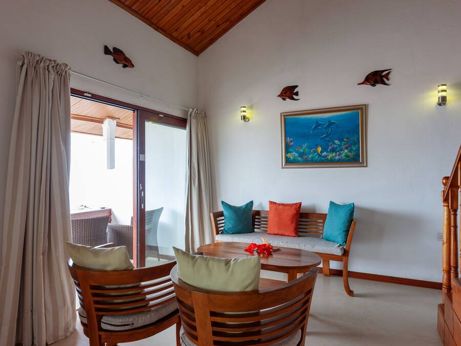 A room with a wooden dining table, chairs, a sofa with colorful cushions, a painting of fish on the wall, and a staircase.