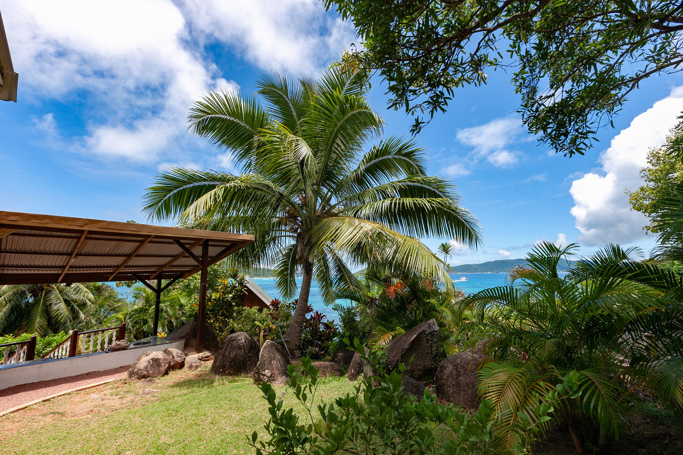 A covered outdoor area with a view of the ocean, palm trees, and a building with a roof.