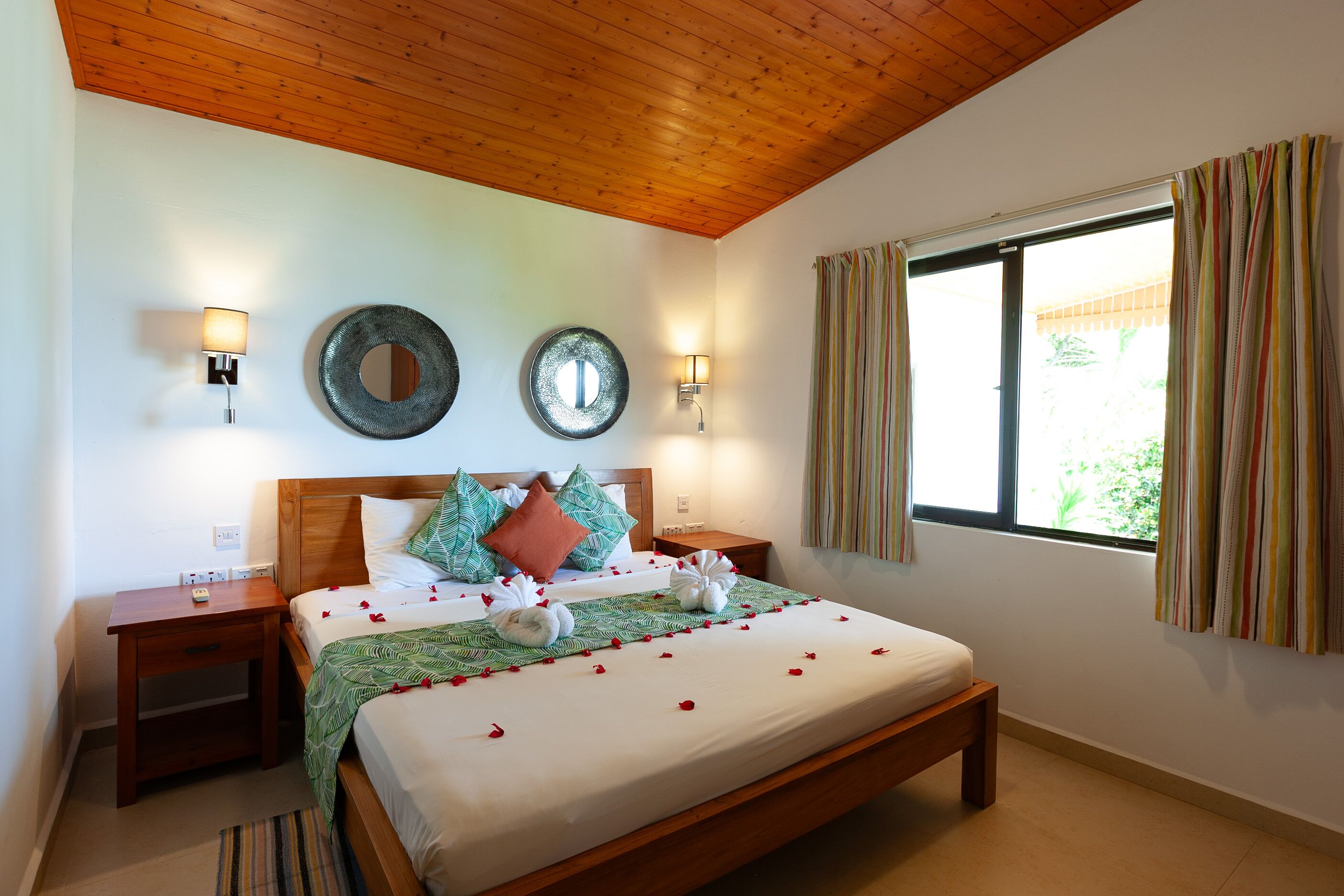 A neatly made bed with pillows and a blanket, two bedside tables with lamps, a large window with curtains, and a wooden ceiling.