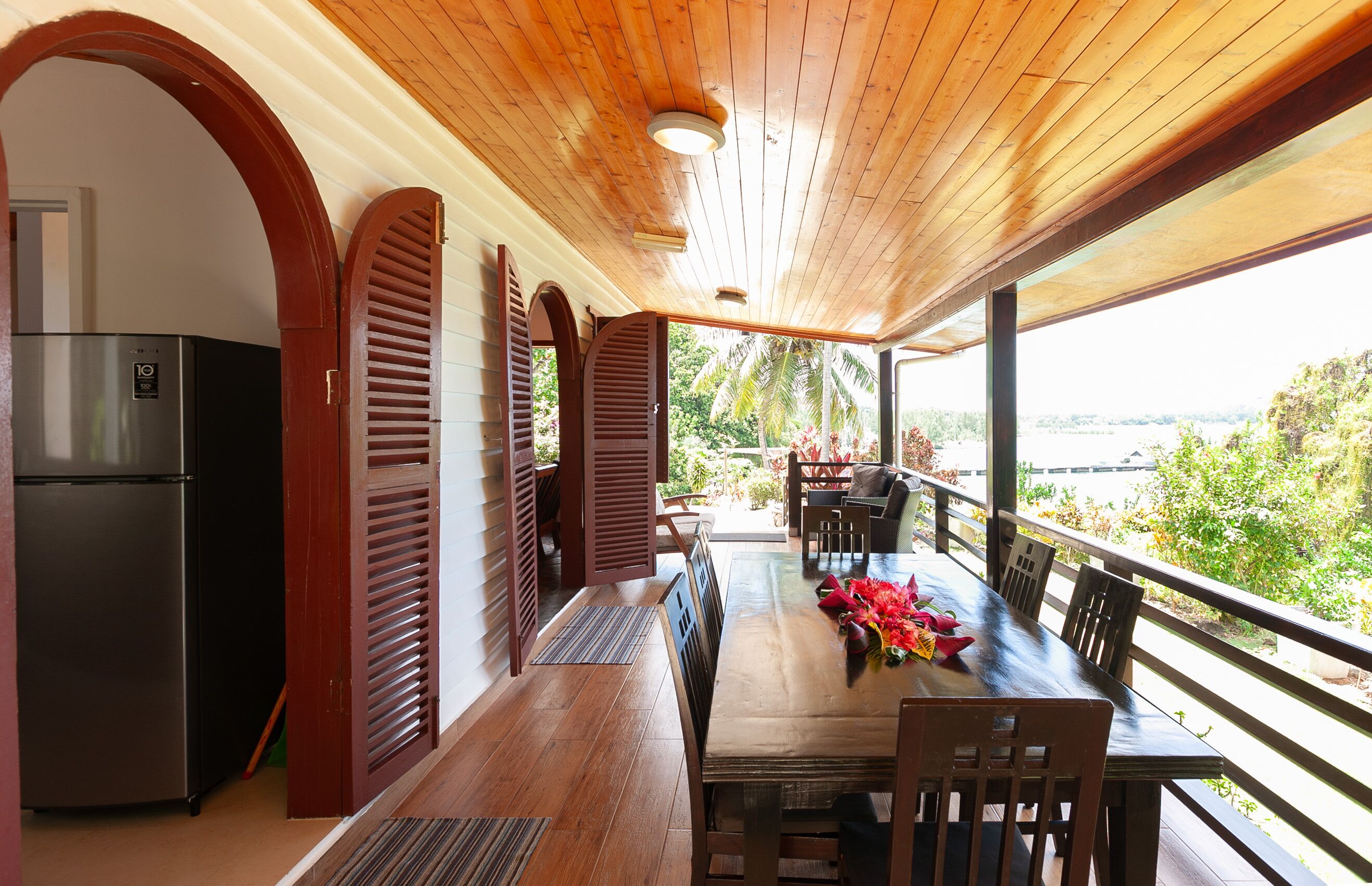 Un espacio de comedor al aire libre con techo, con una mesa y sillas de madera, un refrigerador y vistas al exterior.