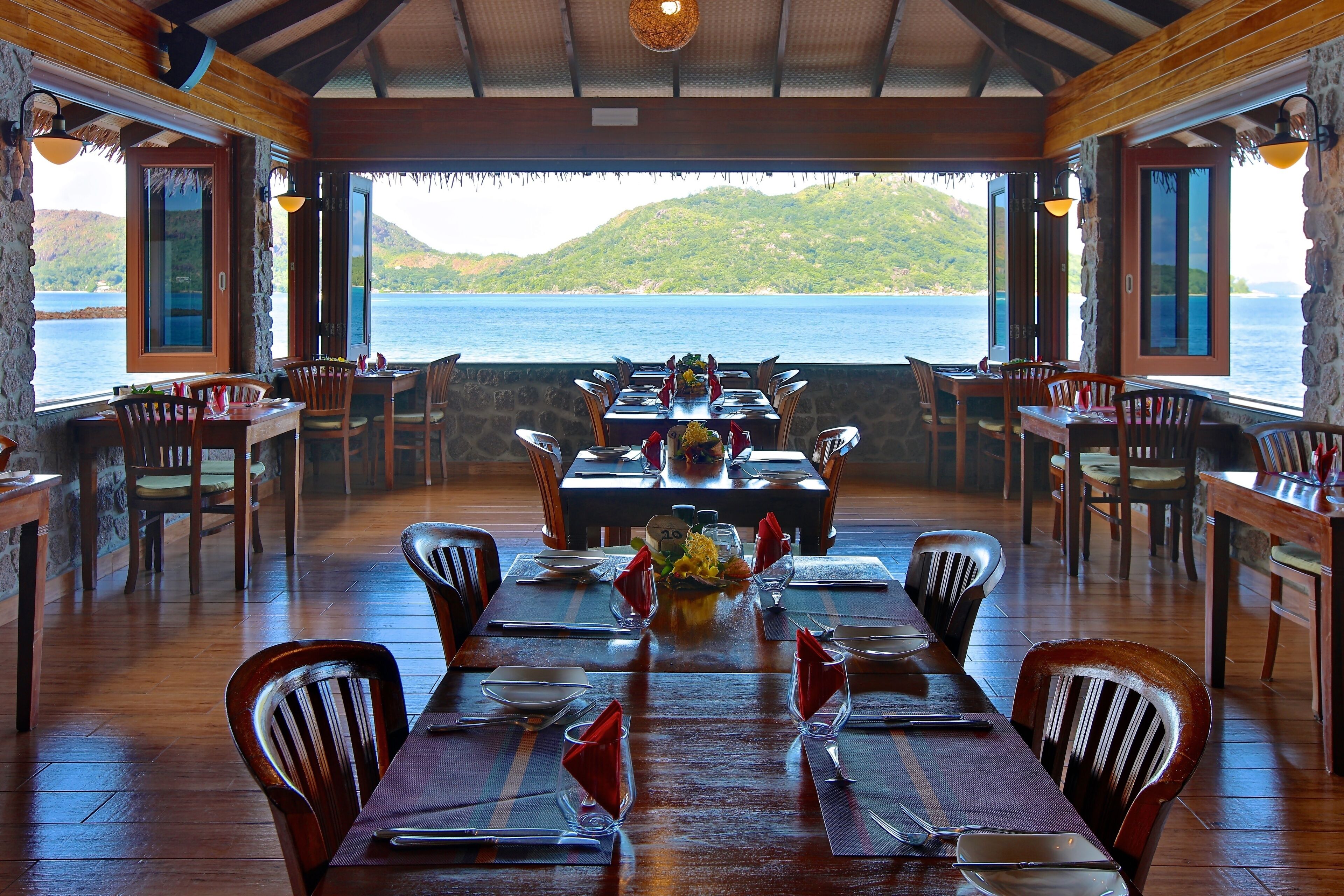 A restaurant with a wooden interior, tables set for dining, and a view of the sea and hills.