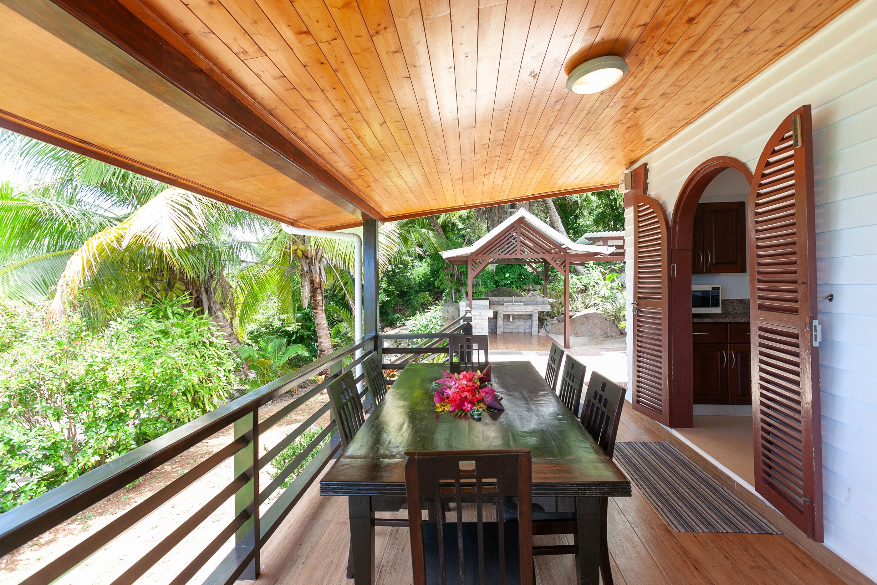 A covered outdoor dining area with a wooden table and chairs, surrounded by lush greenery and a wooden pergola.
