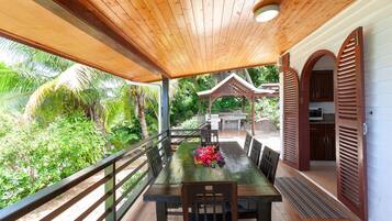 A covered outdoor dining area with a wooden table and chairs, surrounded by lush greenery and a wooden pergola.