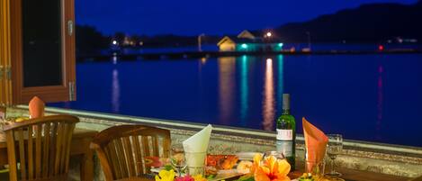 Ein Esszimmerstisch mit Flasche, Gläsern und einem Teller Essen, mit Blick auf ruhiges Wasser in der Nacht.