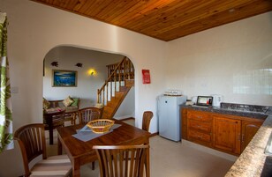 Uma área de refeições com mesa e cadeiras de madeira, uma escada que leva a outro andar, uma cozinha com geladeira e micro-ondas, e um quadro na parede.