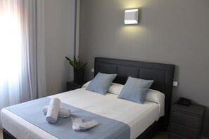 Ein ordentlich bezogenes Bett mit blauer Bettdecke, zwei Kissen und zusammengerollten Handtüchern auf einer weißen Matratze.
