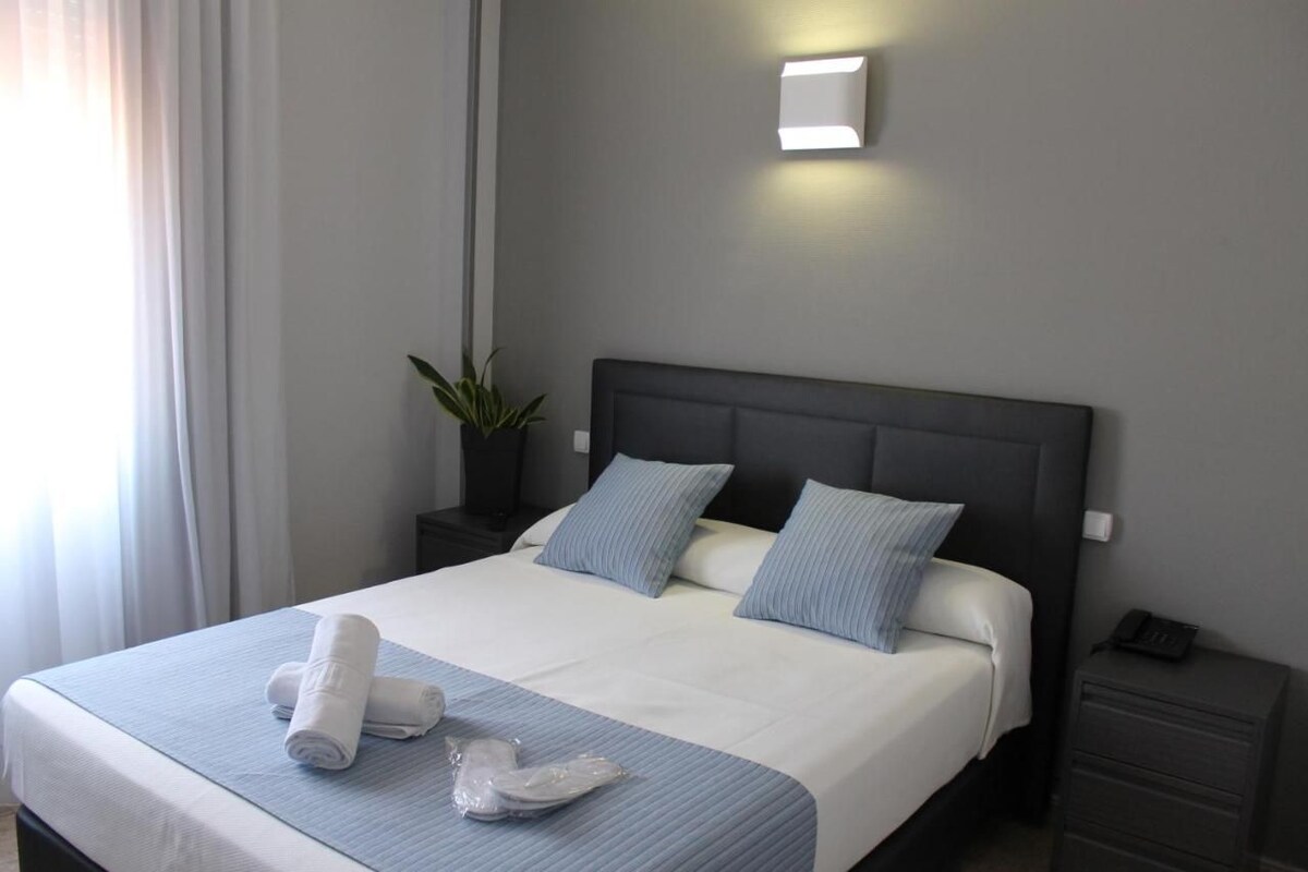 Een net opgemaakt bed met hoofdbord, nachtkastje met lamp en een potplant.