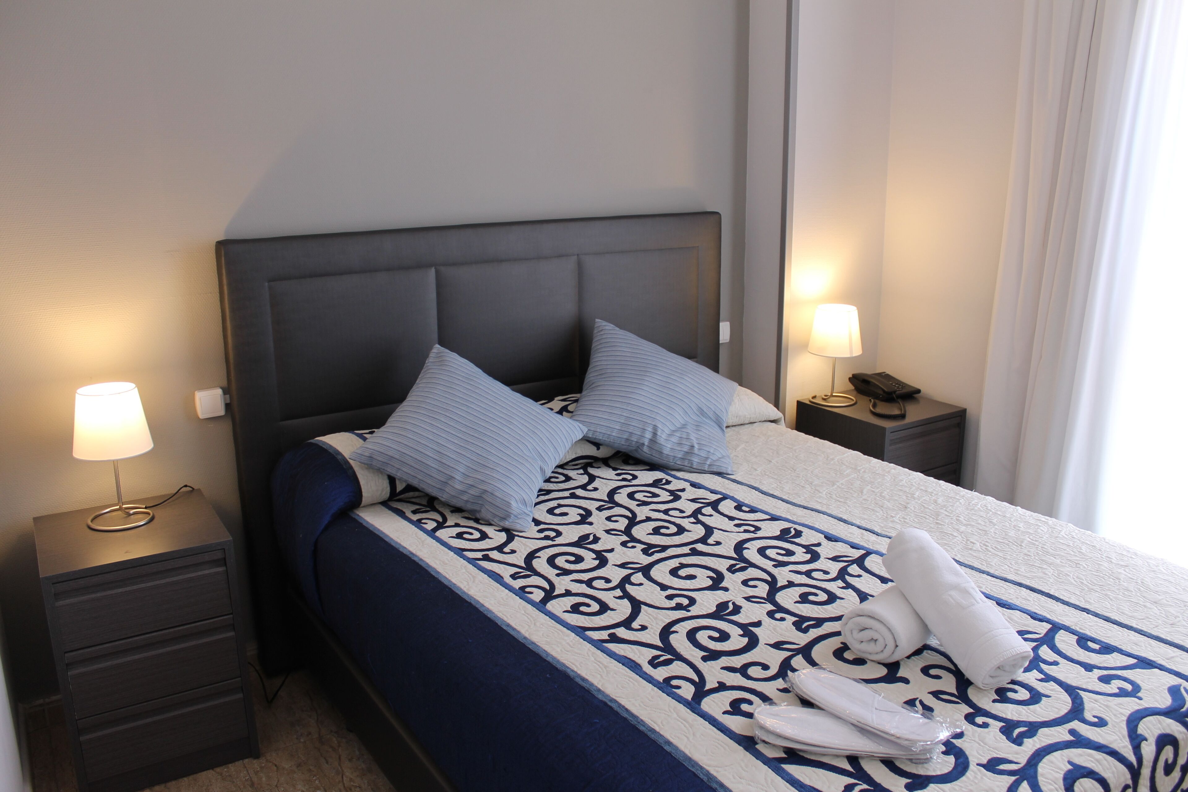 A neatly made bed with a patterned bedspread, two pillows, a bedside table with a lamp, and a folded towel on the bed.