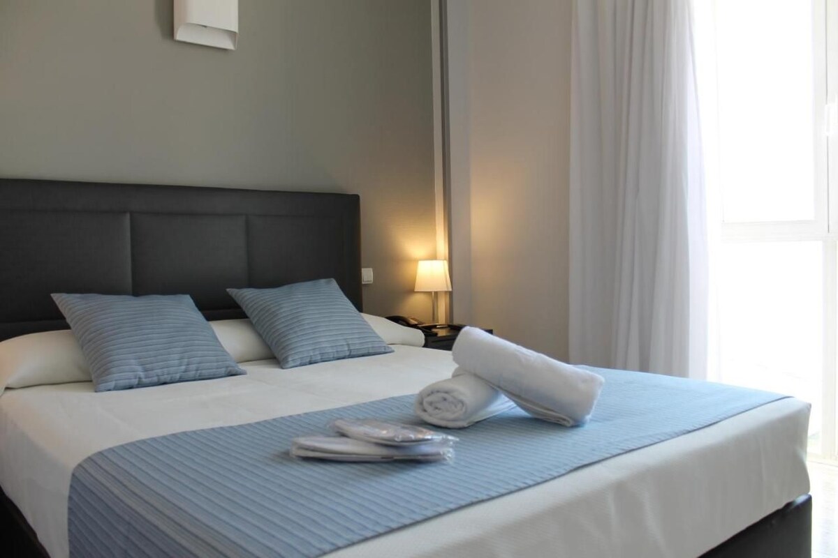 Een net opgemaakt bed met een blauw beddegoed en kussens, een bedlampje en een raam met gordijnen.