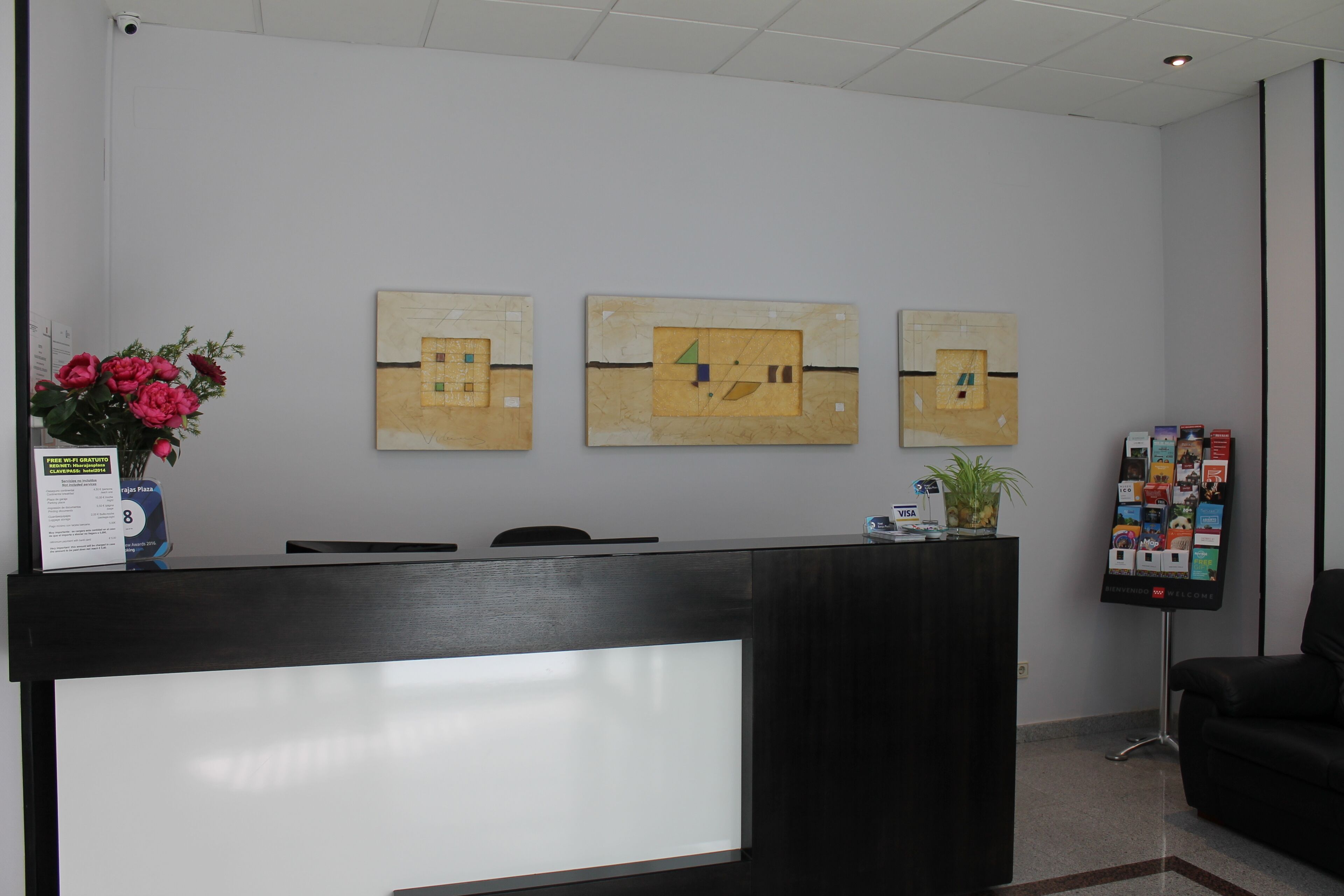 A reception desk with a digital screen, three framed artworks on the wall, a vase of flowers, and a bulletin board with various items.