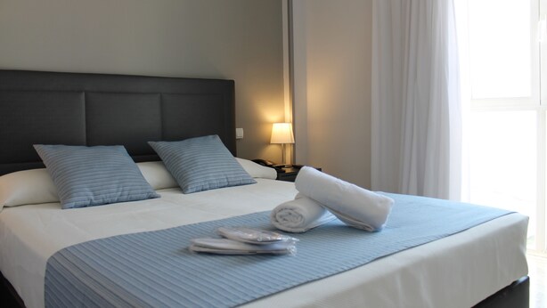 Een net opgemaakt bed met een blauw laken, twee kussens en een opgerolde handdoek.